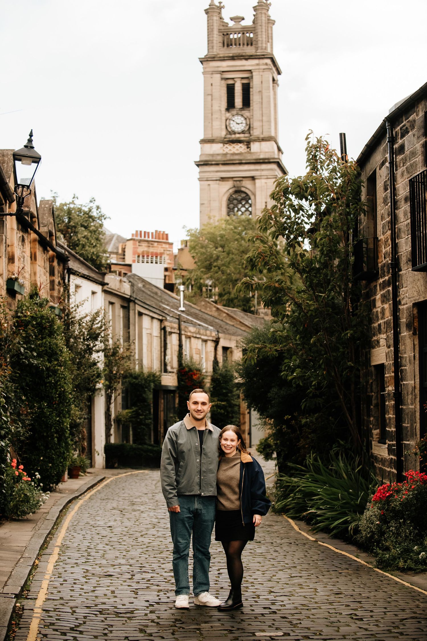 A couple strolls along a charming cobblestone street lined with historic buildings and a church tower.
