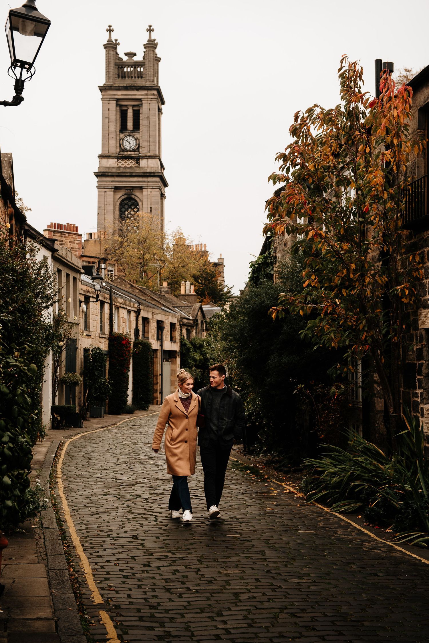People walking along a winding cobblestone street with autumn trees and historic architecture.