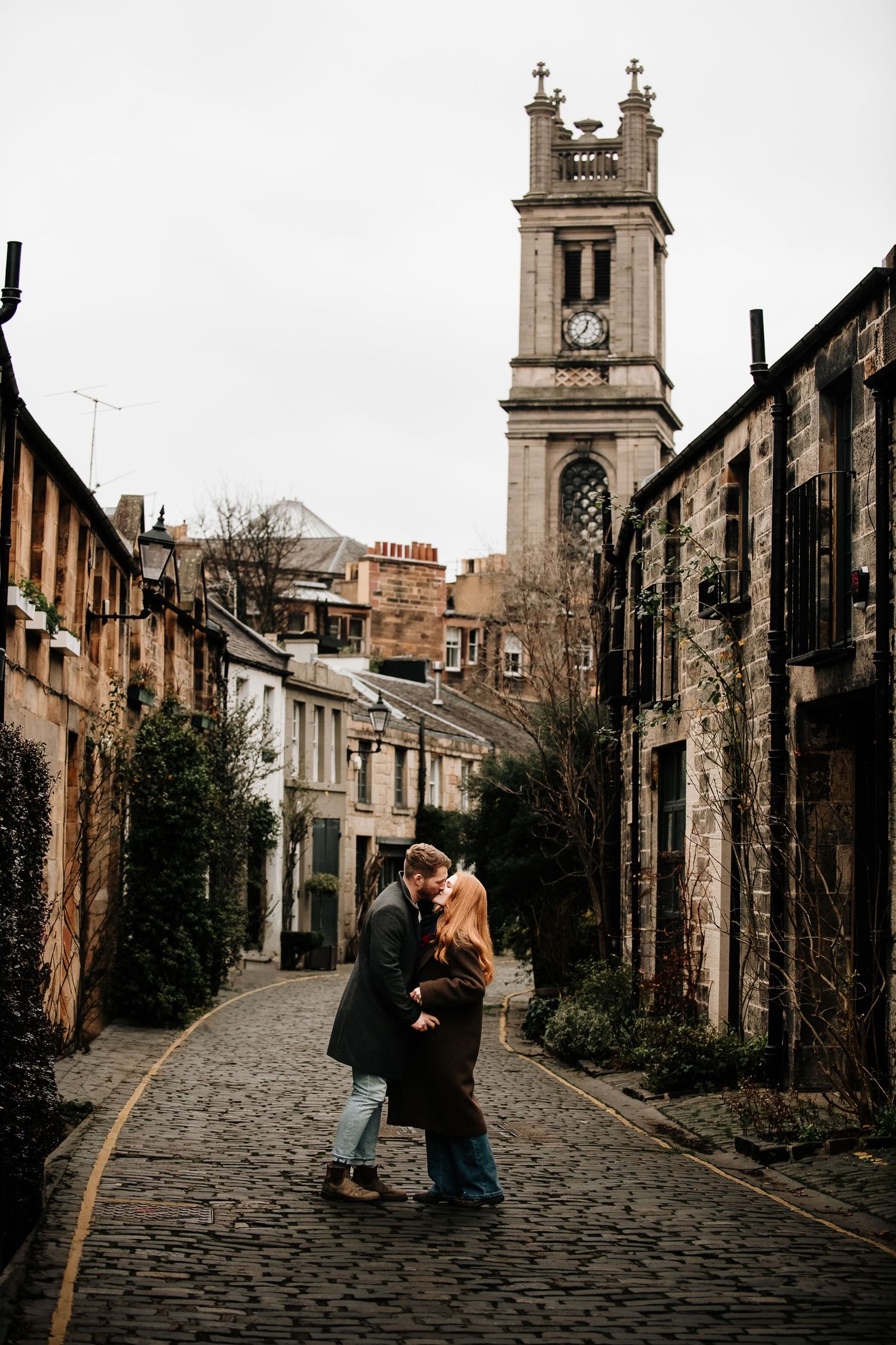 A couple shares an intimate moment on a historic cobblestone street with church spires in the background.