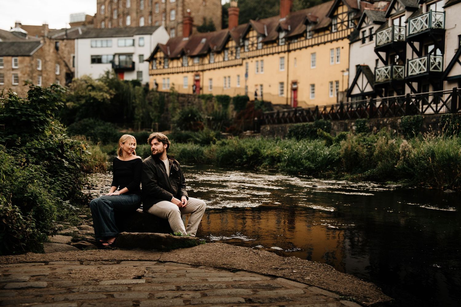 Couple sits along waterway with historic Dean Village buildings and architecture in background.
