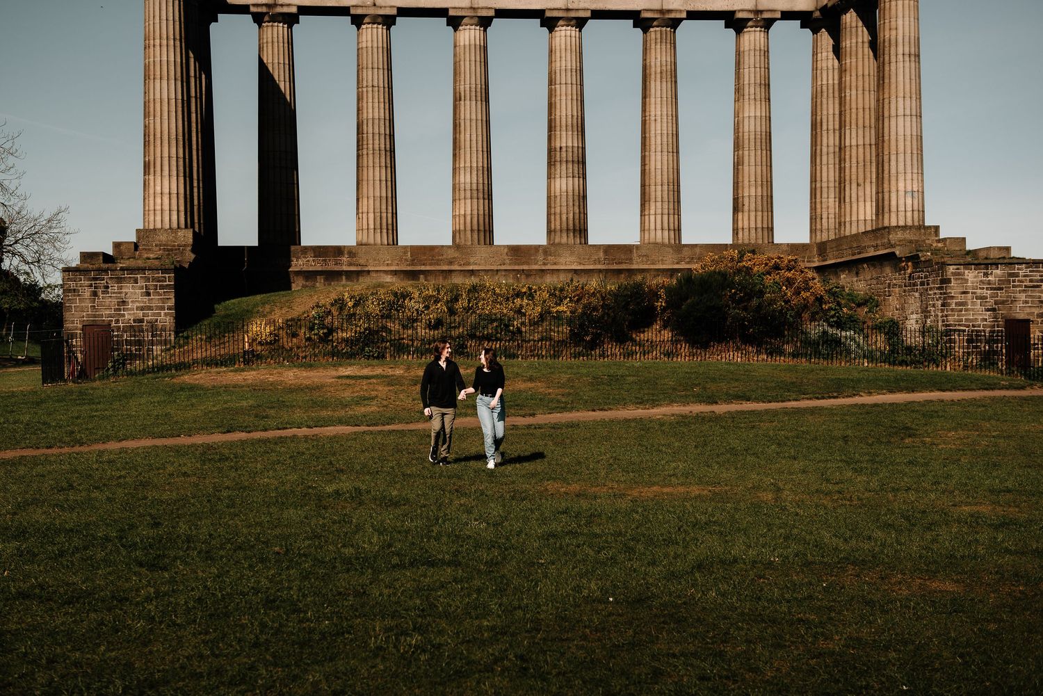 Couple walking on grass in front of ancient Roman-style stone columns at sunset in Edinburgh.