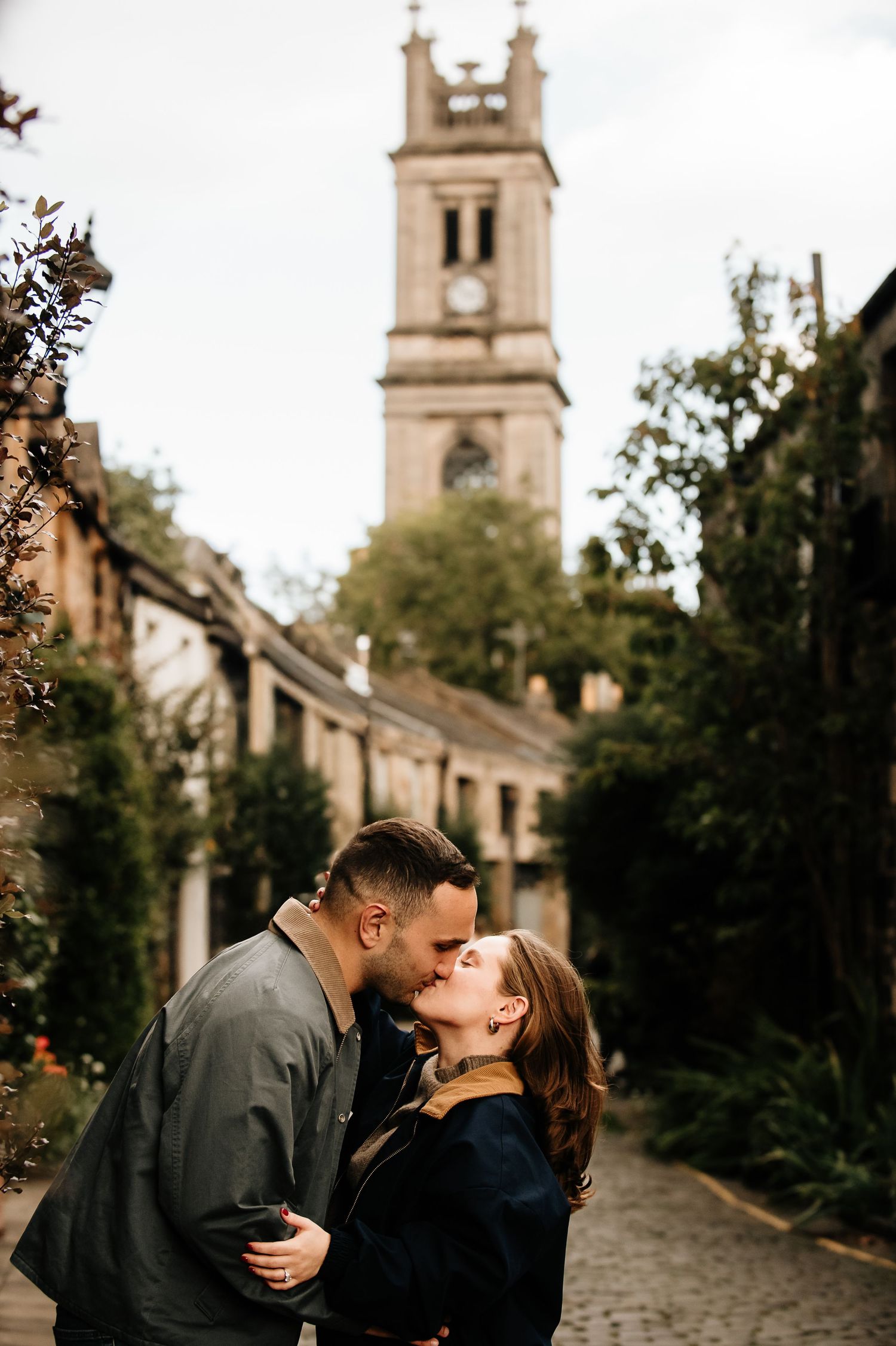 A romantic couple shares an intimate moment on a historic cobblestone street with church spires in the background.
