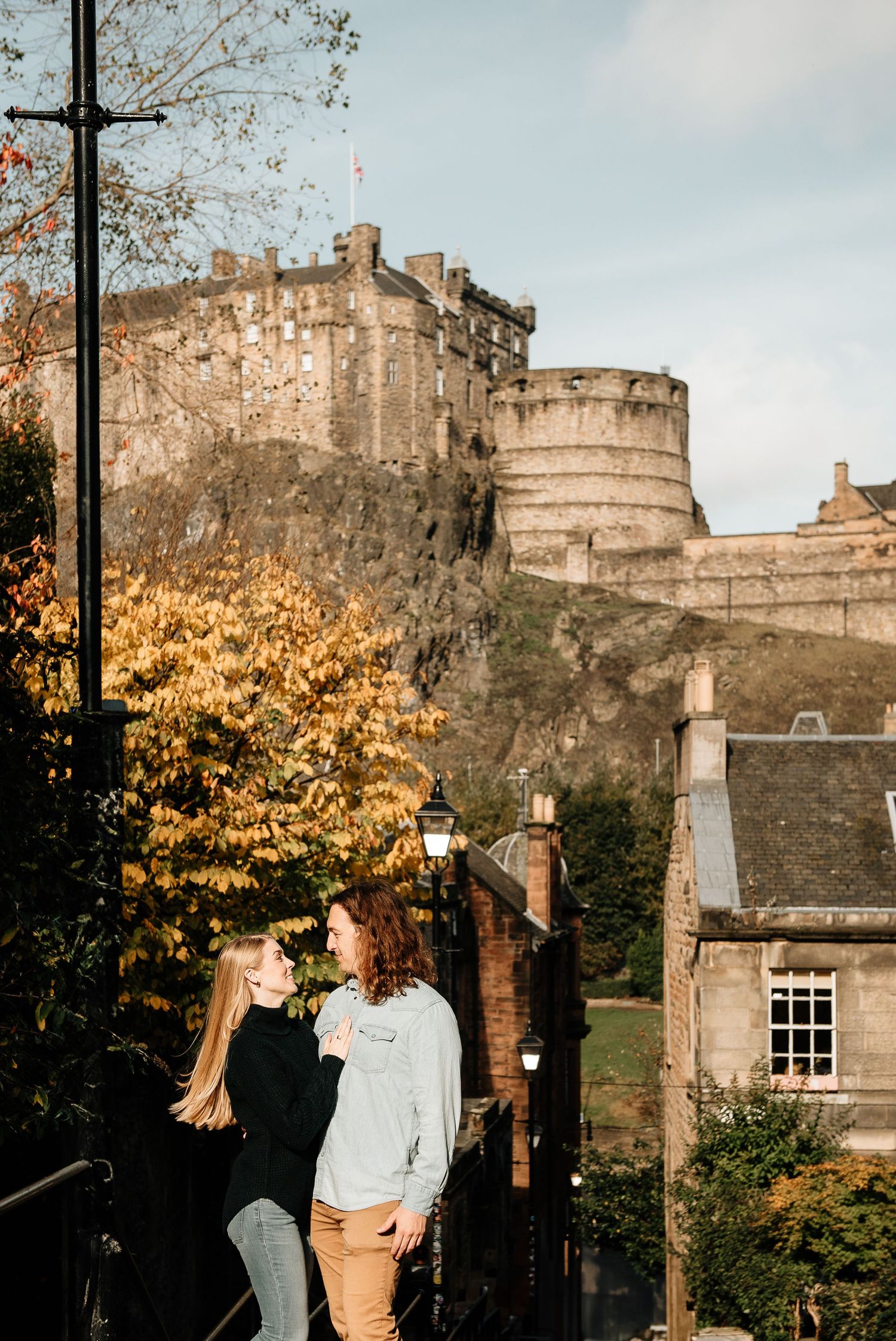 A romantic couple walks along a cobblestone path with Edinburgh Castle looming in the background during autumn.