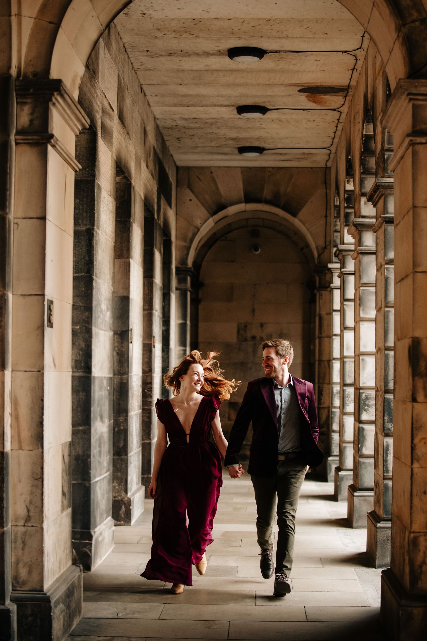 A couple walks through a dramatic stone archway corridor with natural light streaming in from the windows. Edinburgh