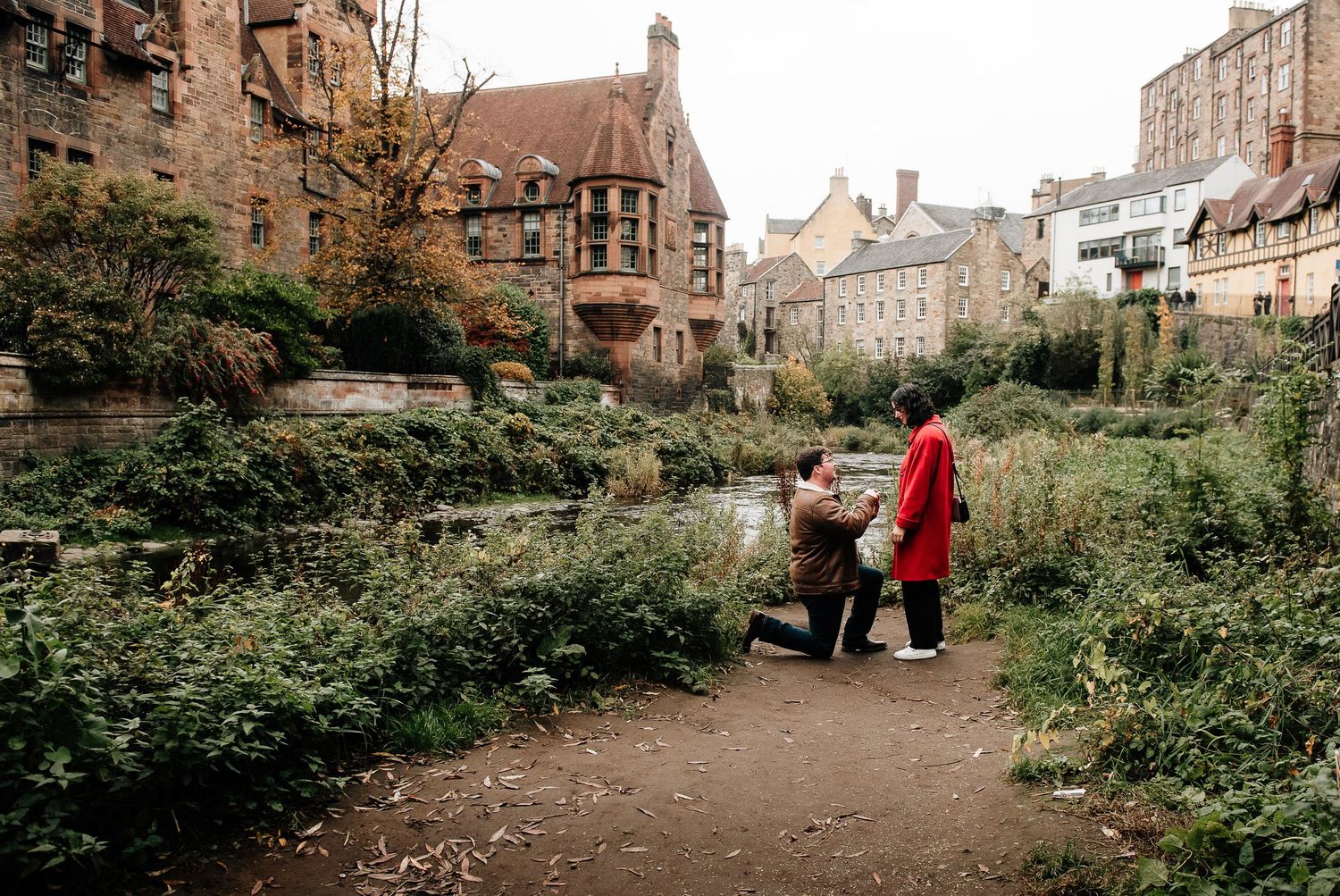 A couple walks through a lush garden path with historic stone buildings in the background.
