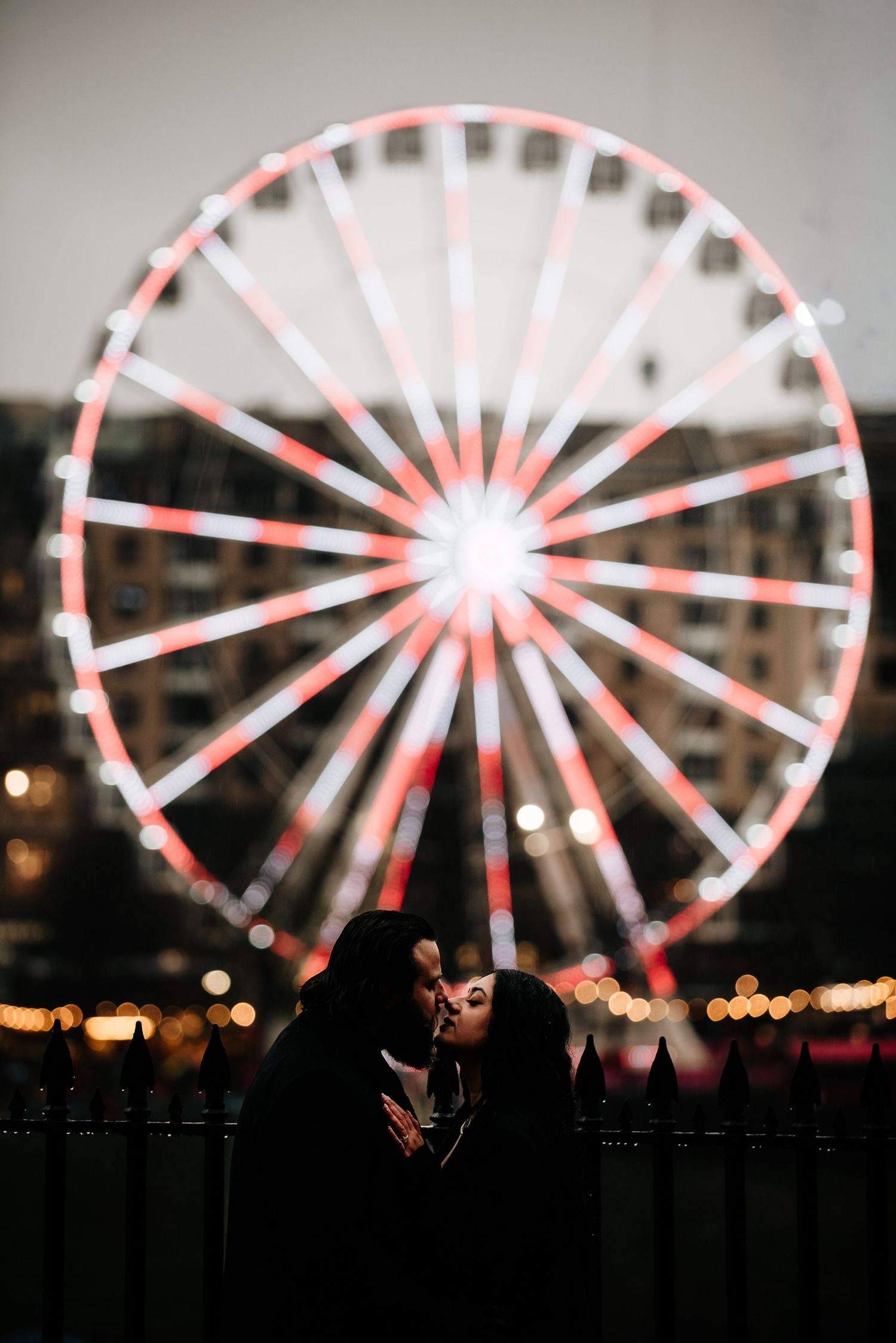 Silhouettes against illuminated ferris wheel at night with red and white lights creating romantic carnival atmosphere.