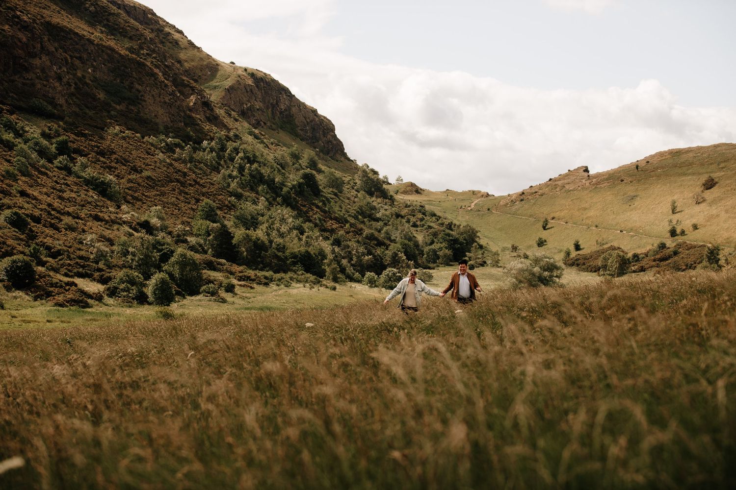Two figures stand in a grassy field with rolling hills and mountains in the distance.
