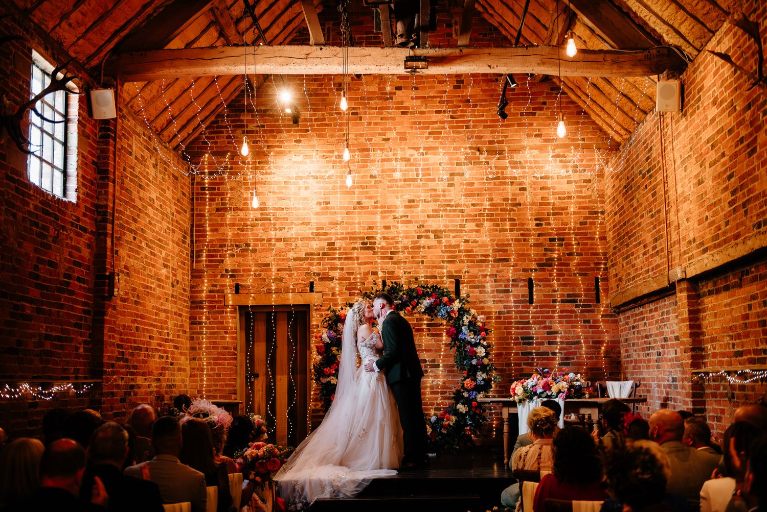 Bride and groom exchange vows at a rustic brick barn wedding ceremony with floral arch and warm Edison bulb lighting.