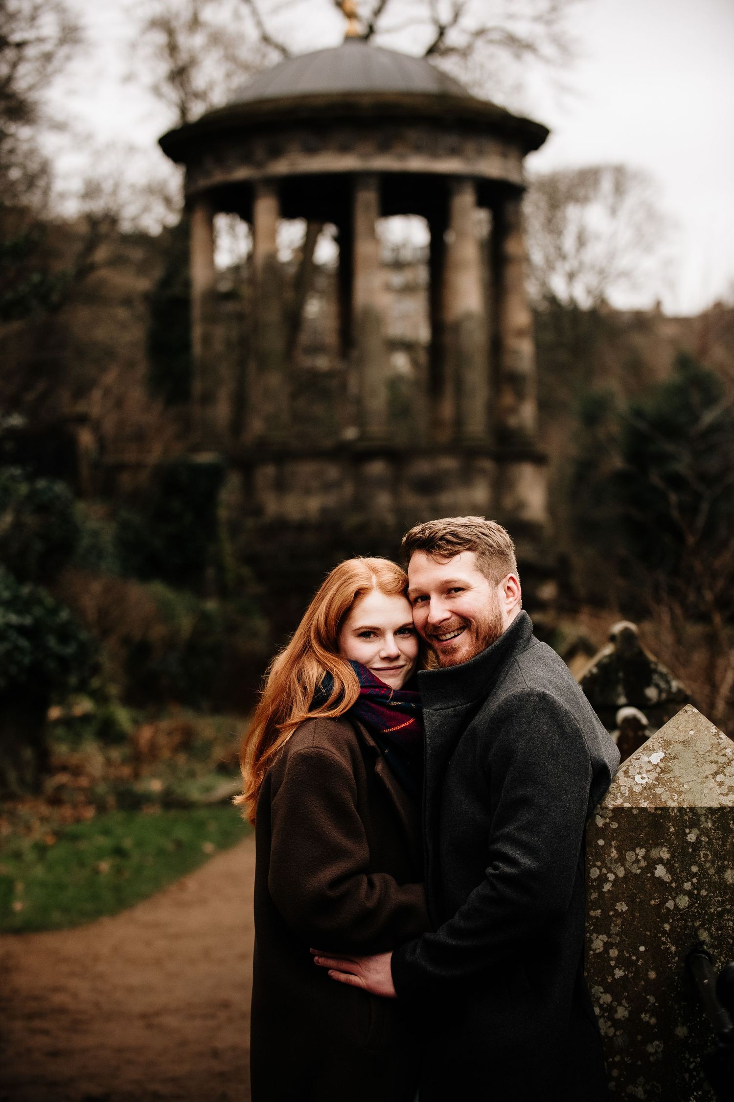 Couple shares intimate moment near historic stone gazebo in autumn park setting.