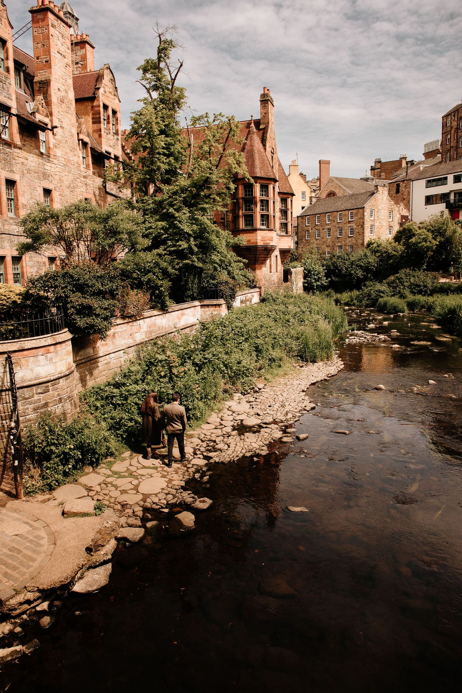 Historic Dean Village in Edinburgh with a person standing by the Water of Leith, surrounded by old stone buildings and lush greenery.