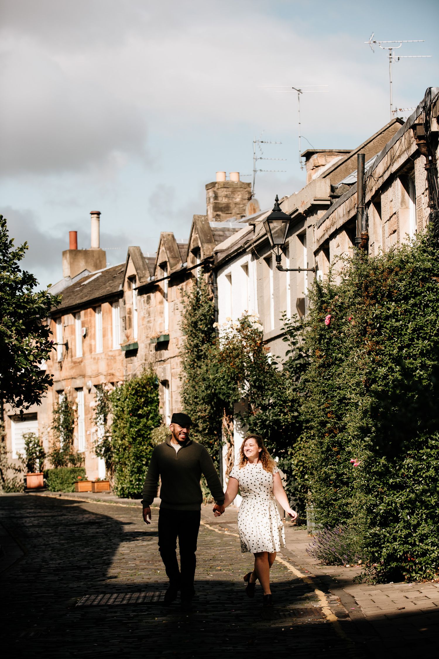 Couple holding hands walking on cobblestone street lined with historic stone buildings and lush greenery on a sunny day.