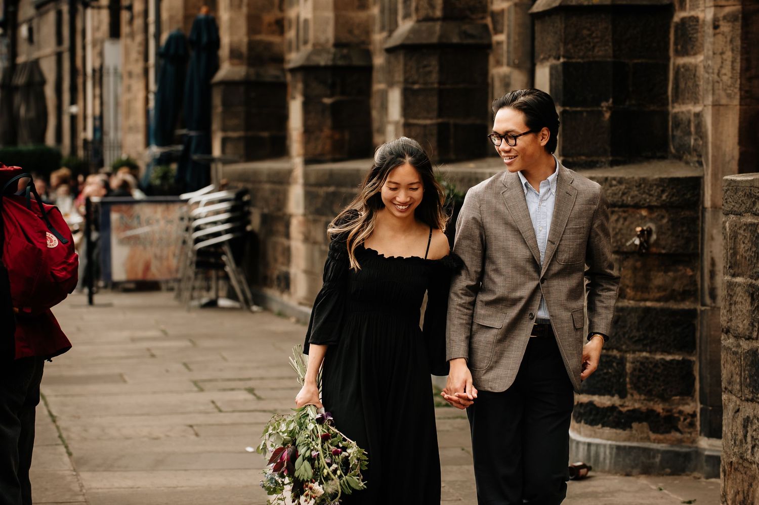 A couple in formal attire walks hand in hand along a historic stone building with a beautiful bouquet of flowers.
