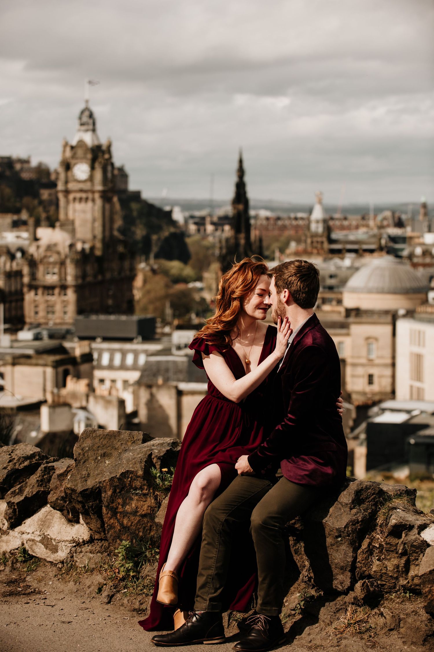 A romantic couple shares an intimate moment overlooking Edinburgh's historic cityscape with the Balmoral Clock Tower visible.