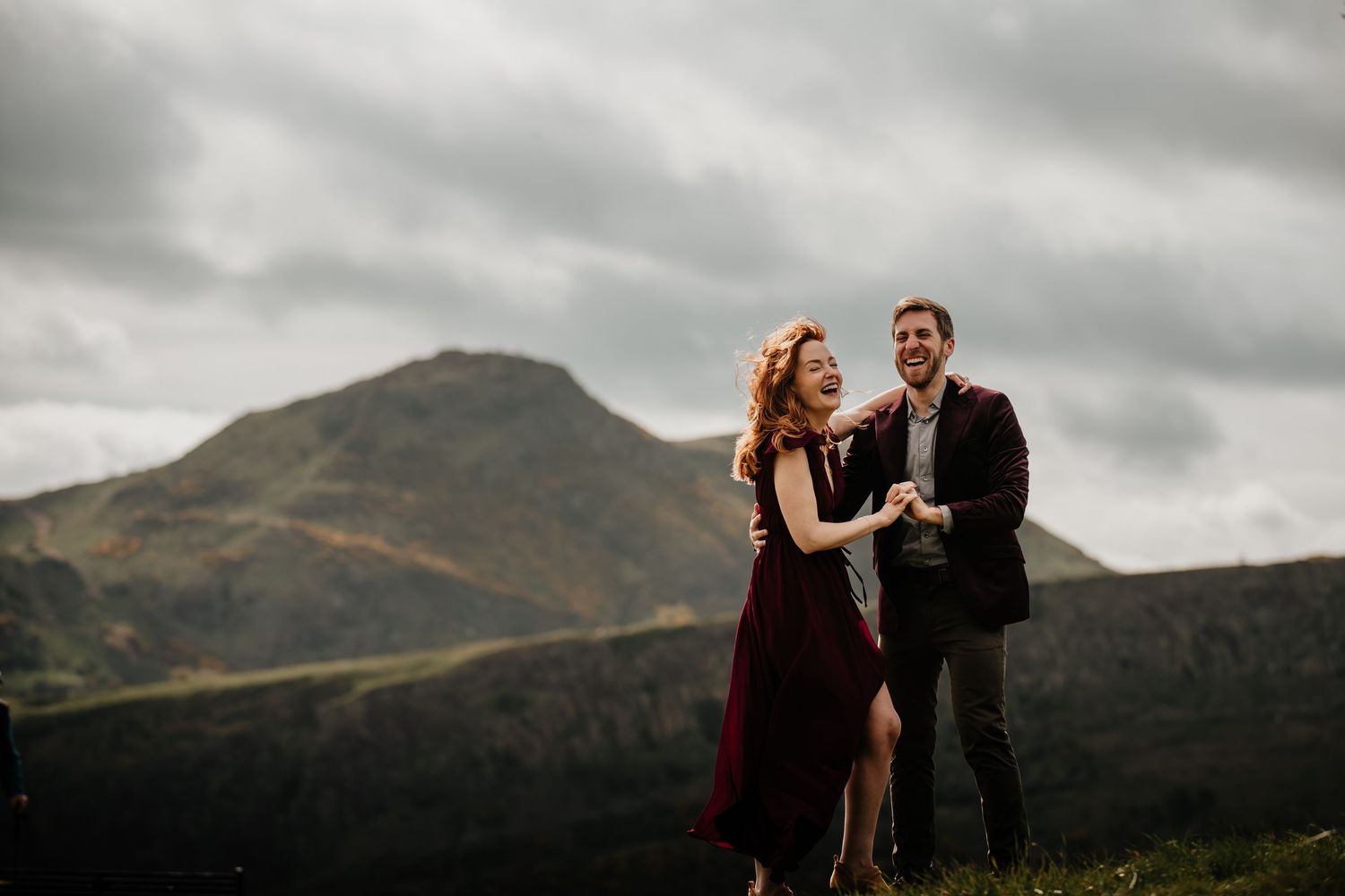 A couple in elegant dark attire share a romantic moment against a dramatic Arthur's Seat under moody clouds.