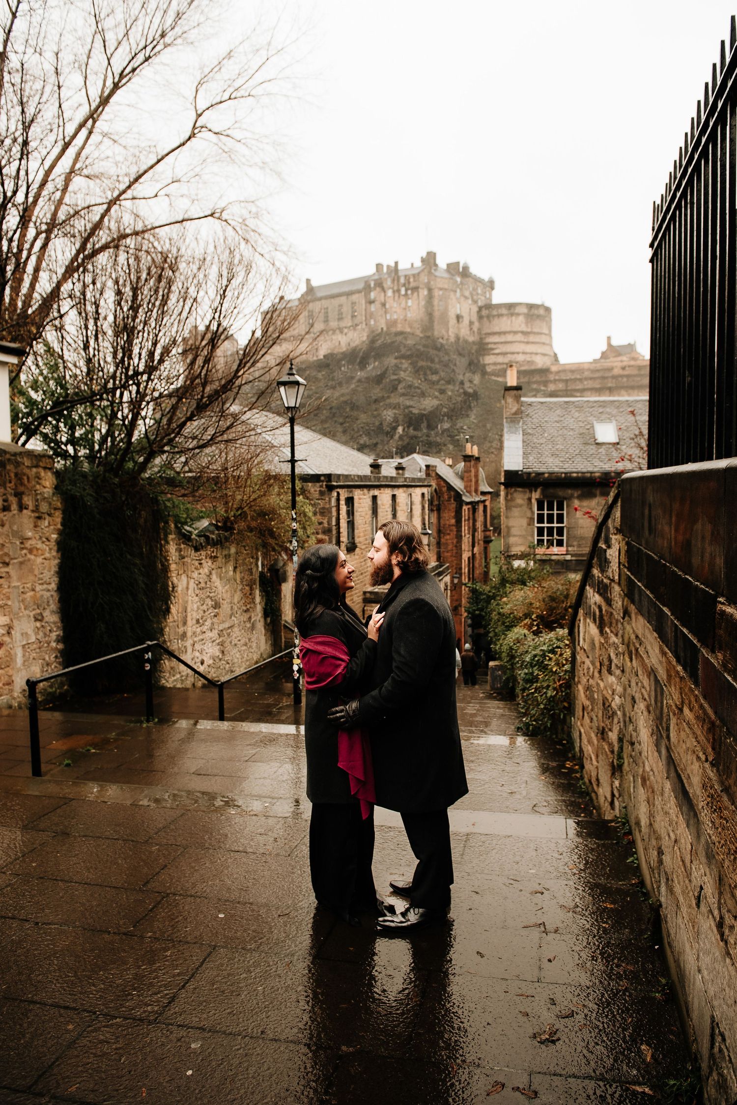 A couple shares an intimate moment on a rainy cobblestone street with Edinburgh Castle looming in the background.
