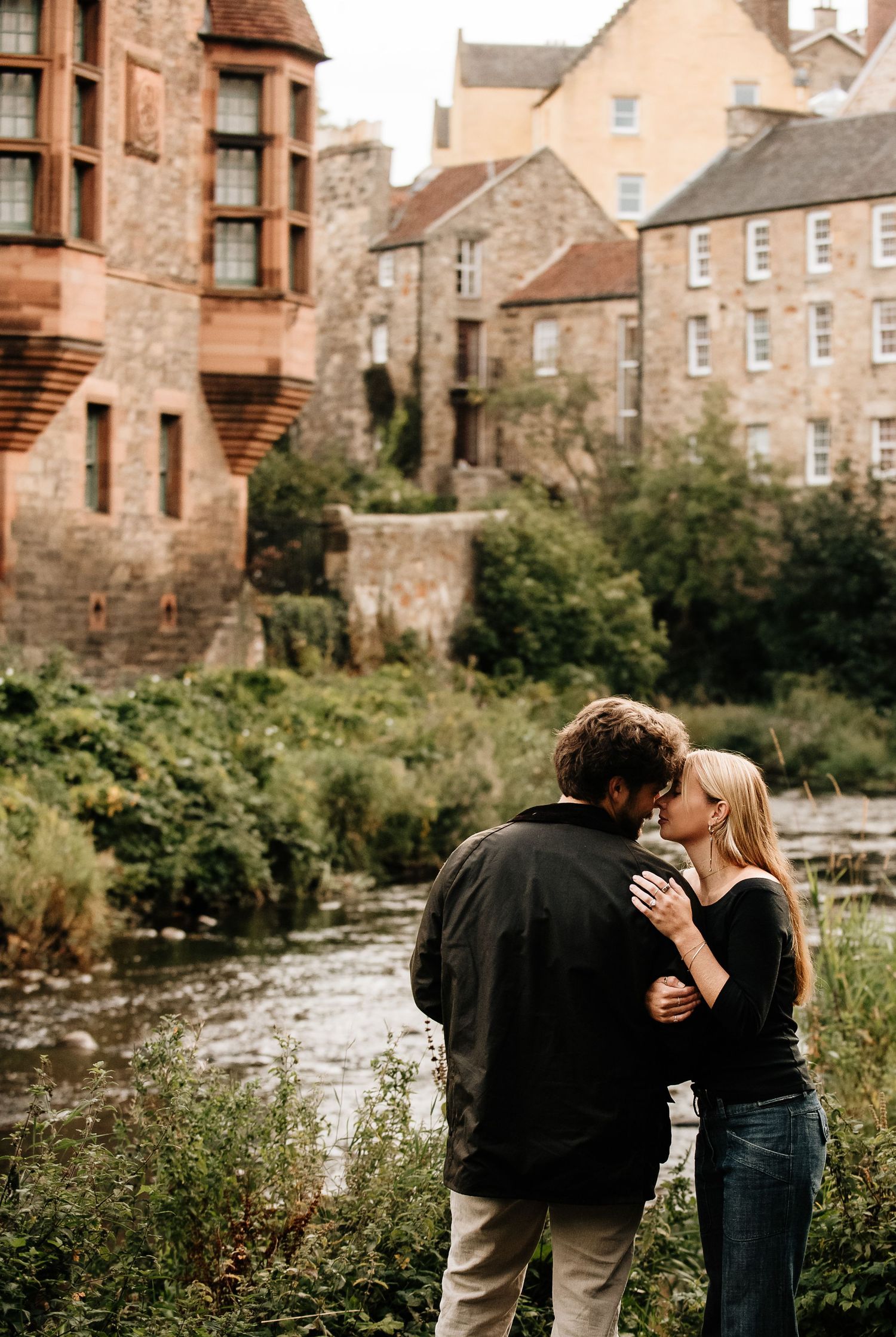 A couple shares tender moments by a scenic waterway with historic stone buildings in the background.