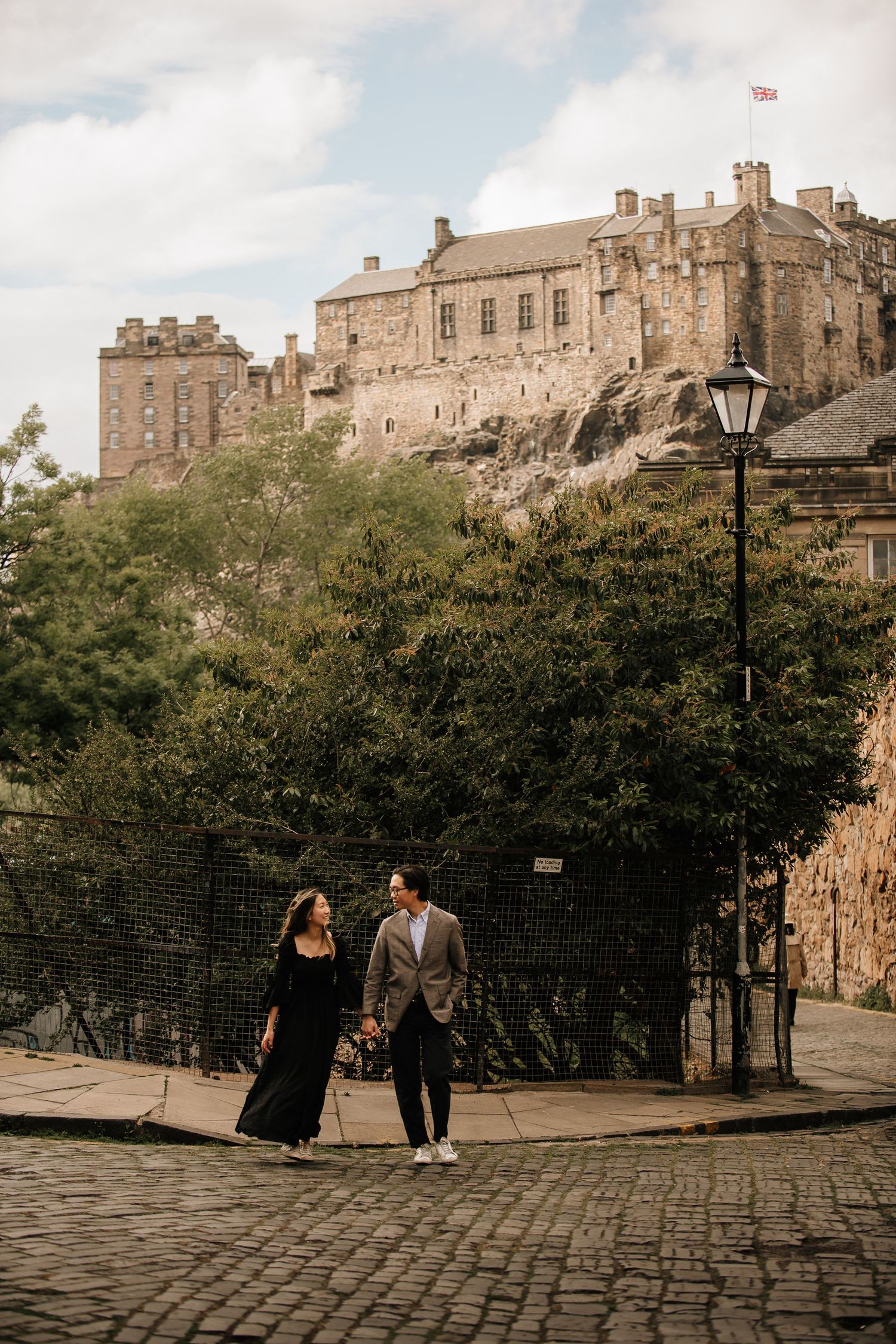 A couple walks along cobblestone paths beneath the majestic Edinburgh Castle on a bright day.