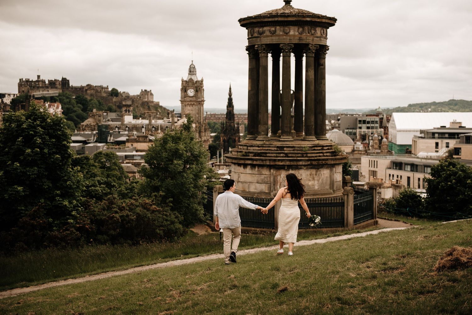 Couple walking hand in hand near Dugald Stewart Monument with Edinburgh skyline behind.