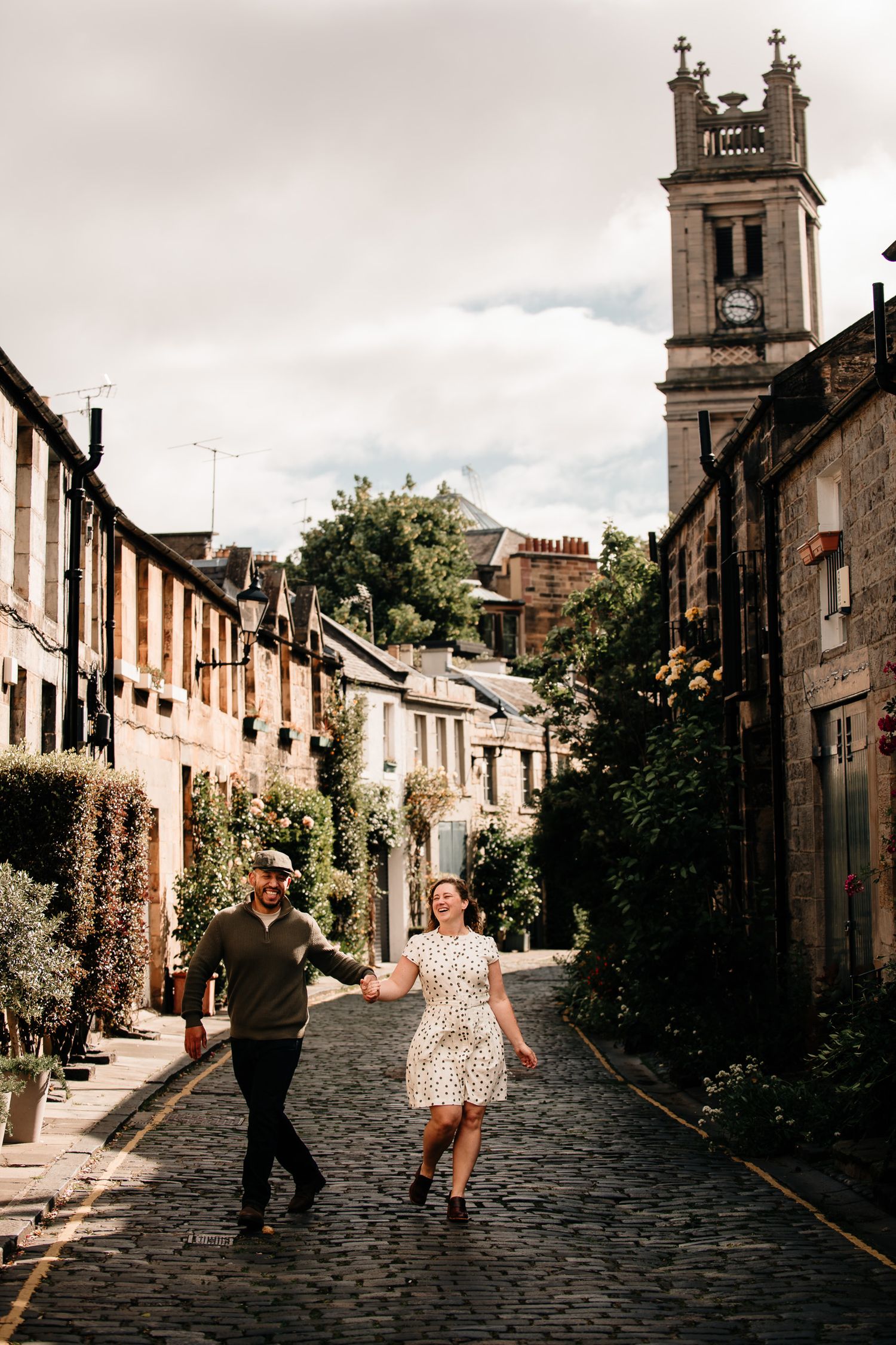 Couple holding hands and walking on cobblestone street in Edinburgh with historic clock tower and charming row houses in background.