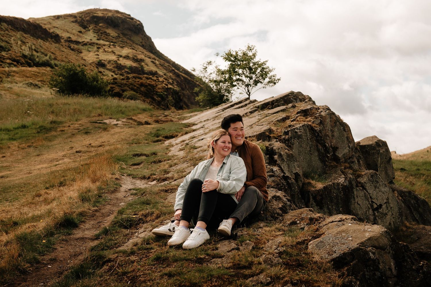 A couple sits together on rocky terrain with a mountainous backdrop.