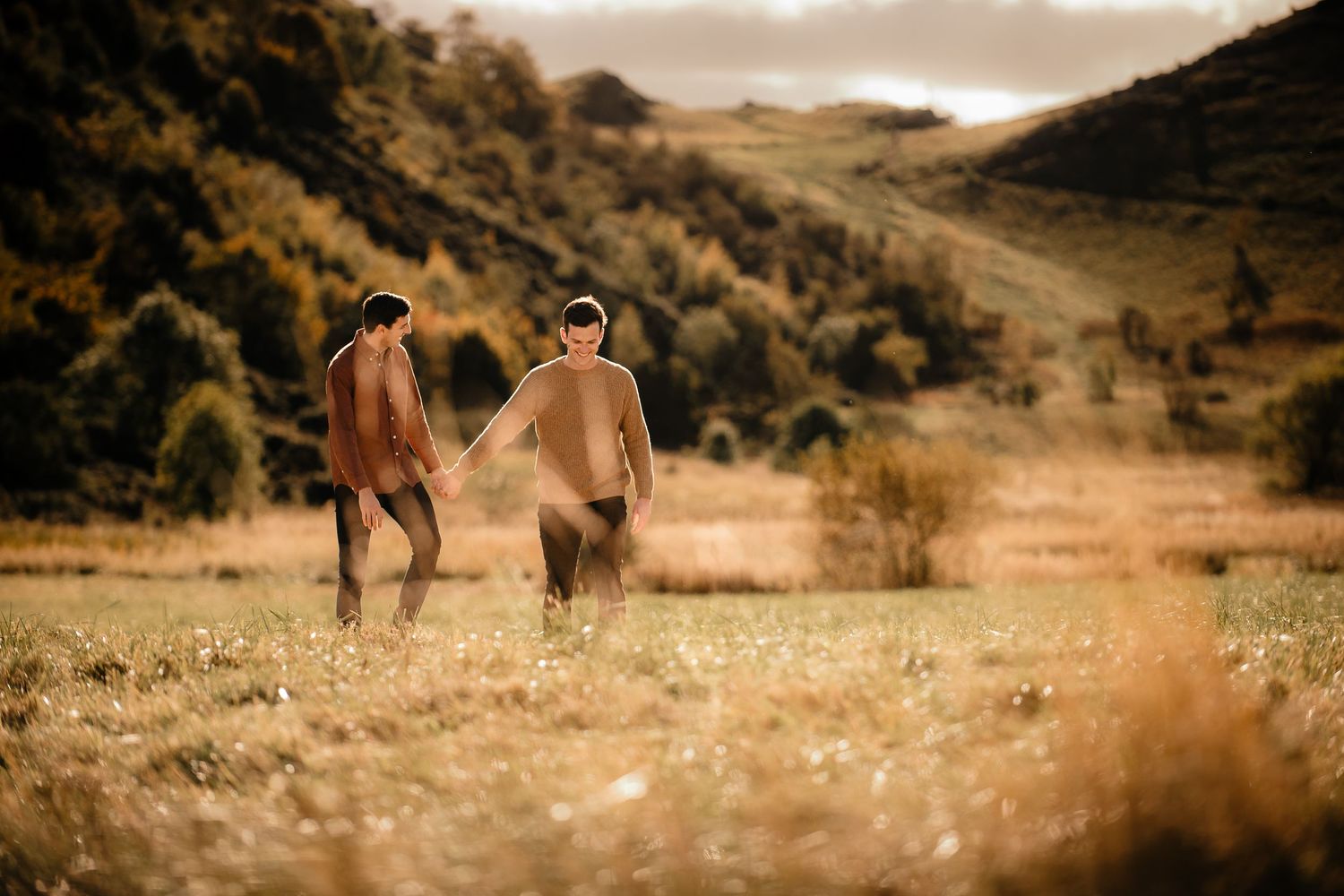 Two people walk through golden grass fields with mountains in warm sunset light.