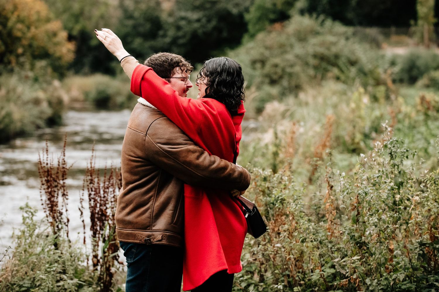 A couple embraces in a natural setting with autumn foliage and water in the background.