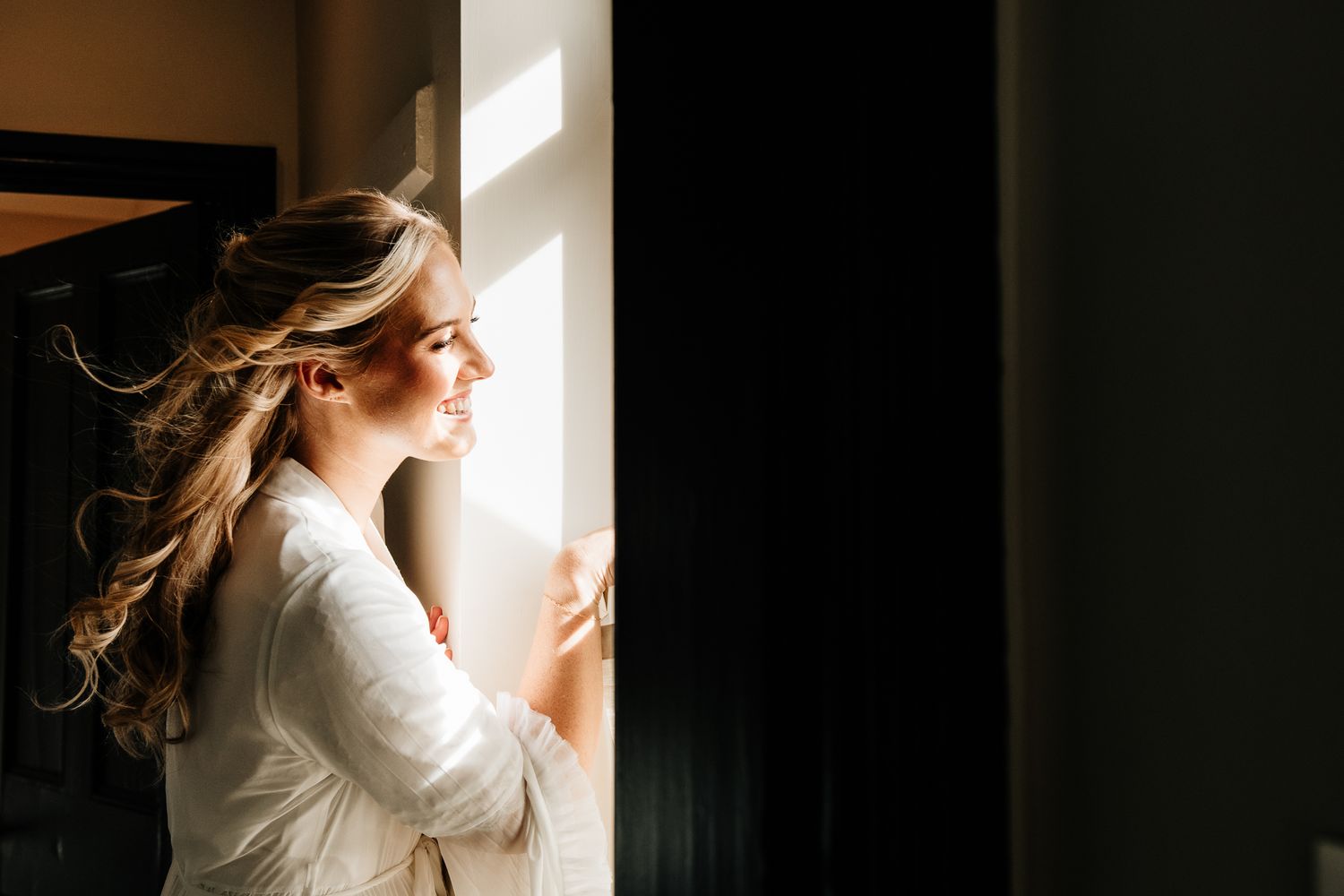 Bride in white robe smiling by sunlit window with elegant curled hair on her wedding day.