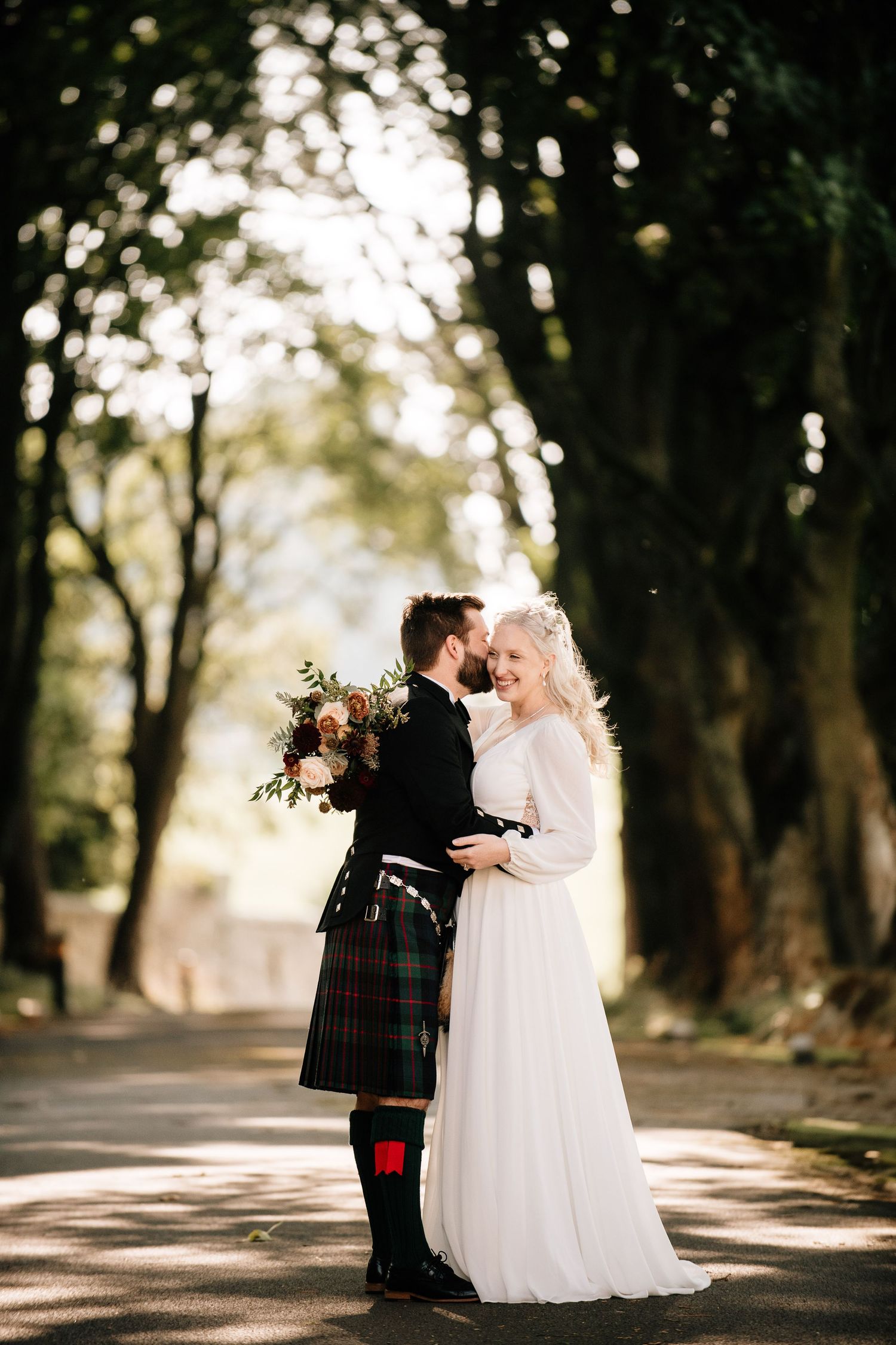 Wedding couple in traditional Scottish attire poses on tree-lined path.