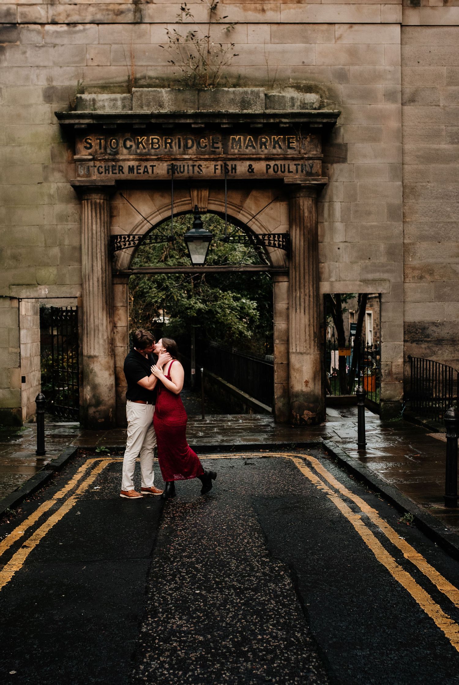 A couple embraces under an ornate stone archway on a wet Edinburgh street.