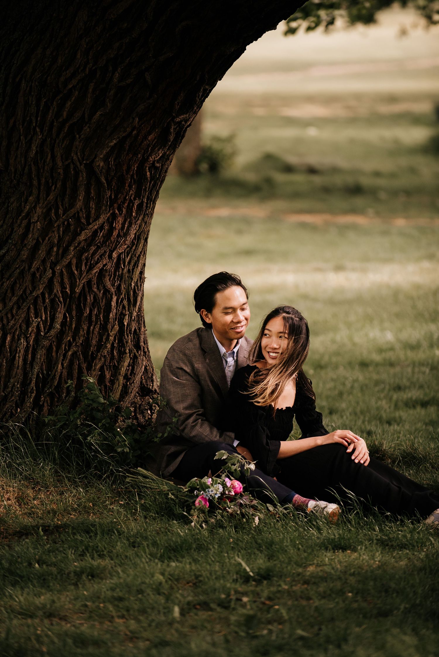A couple shares a joyful moment together while lying in the grass beneath a large tree during a photo session.