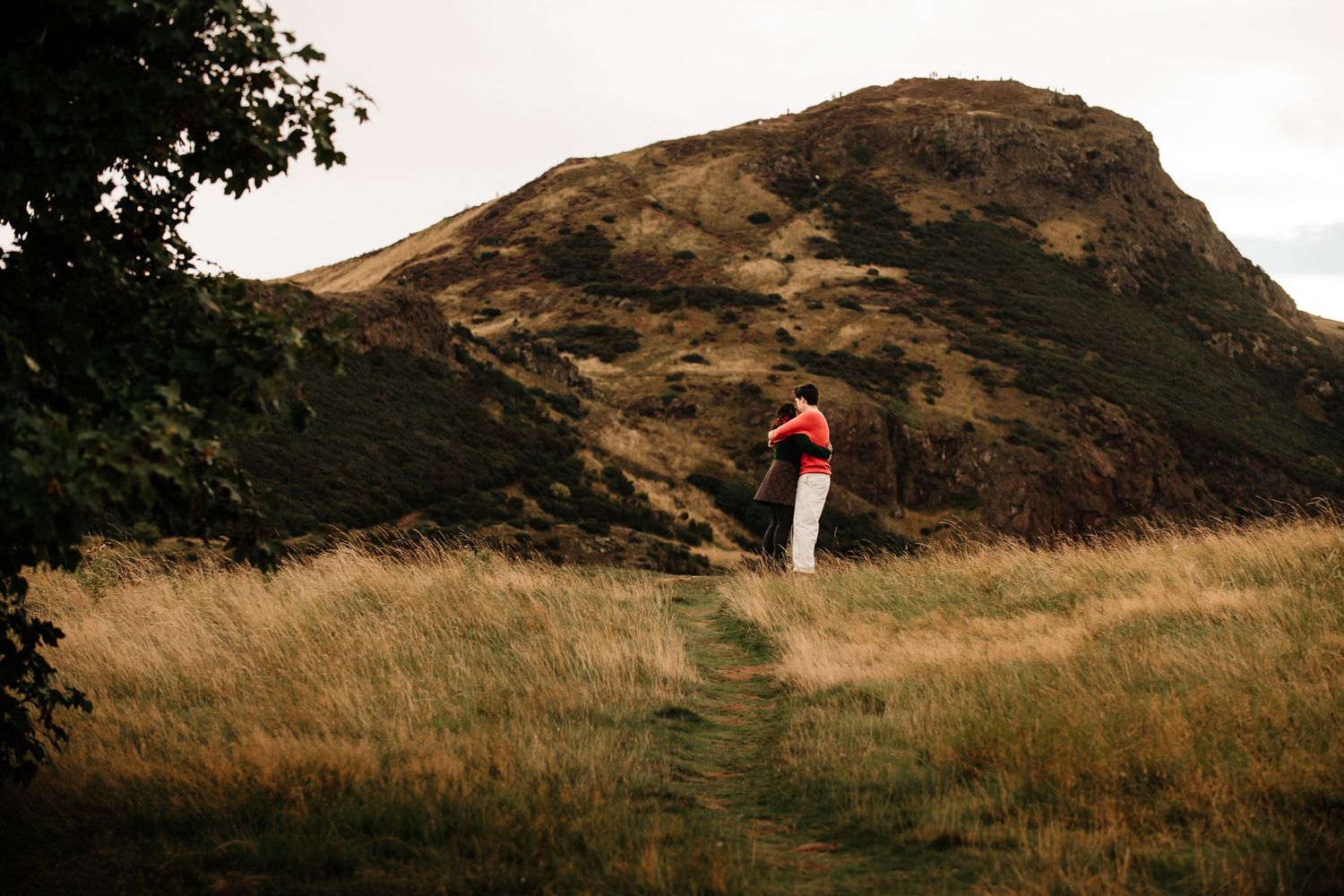 Person in red jacket standing on a grassy trail with a dramatic rocky hill in the background.