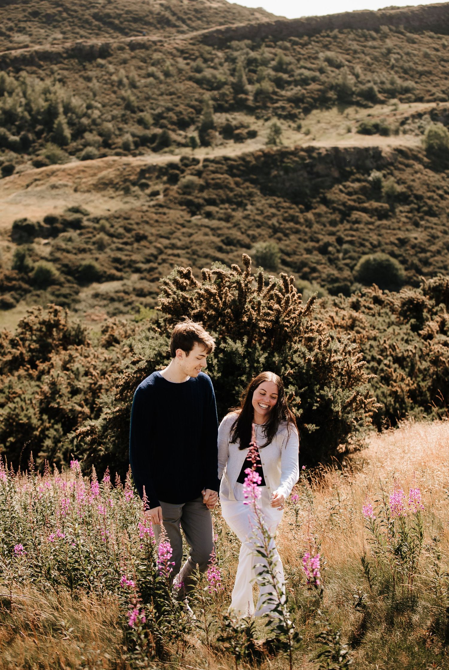 A couple walks through wildflowers and tall grass on a mountainside trail.