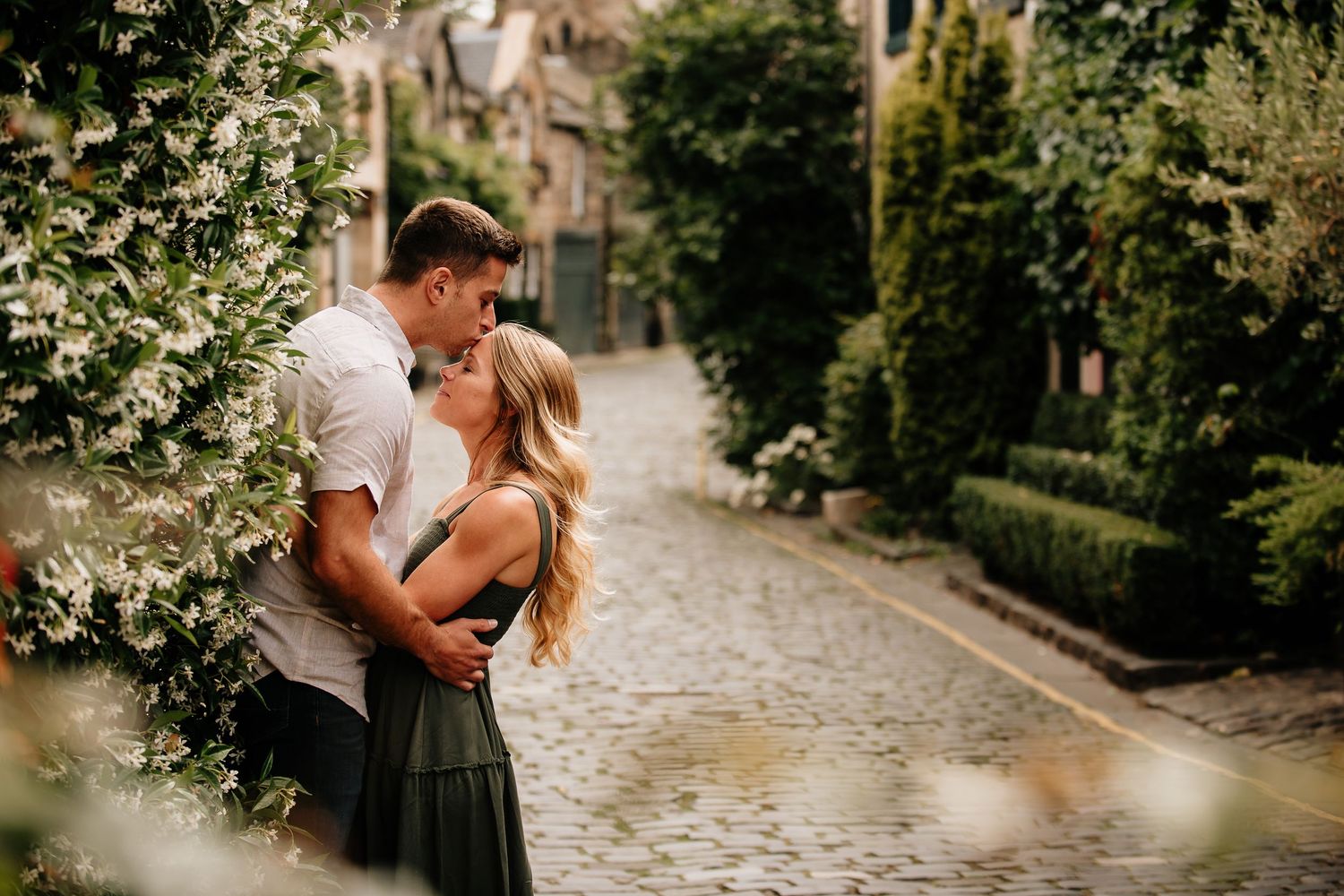 A romantic couple embraces on a cobblestone path surrounded by lush greenery and sunlit trees.