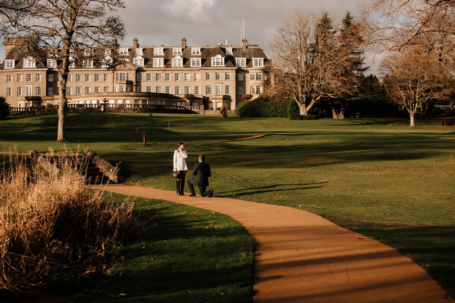 Stone buildings and autumn trees line a scenic path at a historic campus or estate.