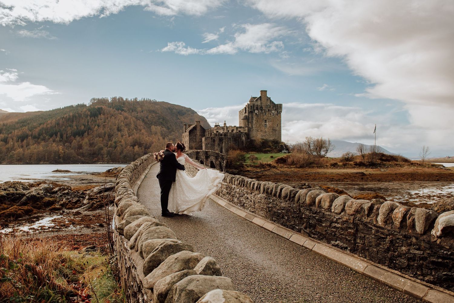 Panoramic view of Eilean Donan Castle with wedding couple on stone bridge.