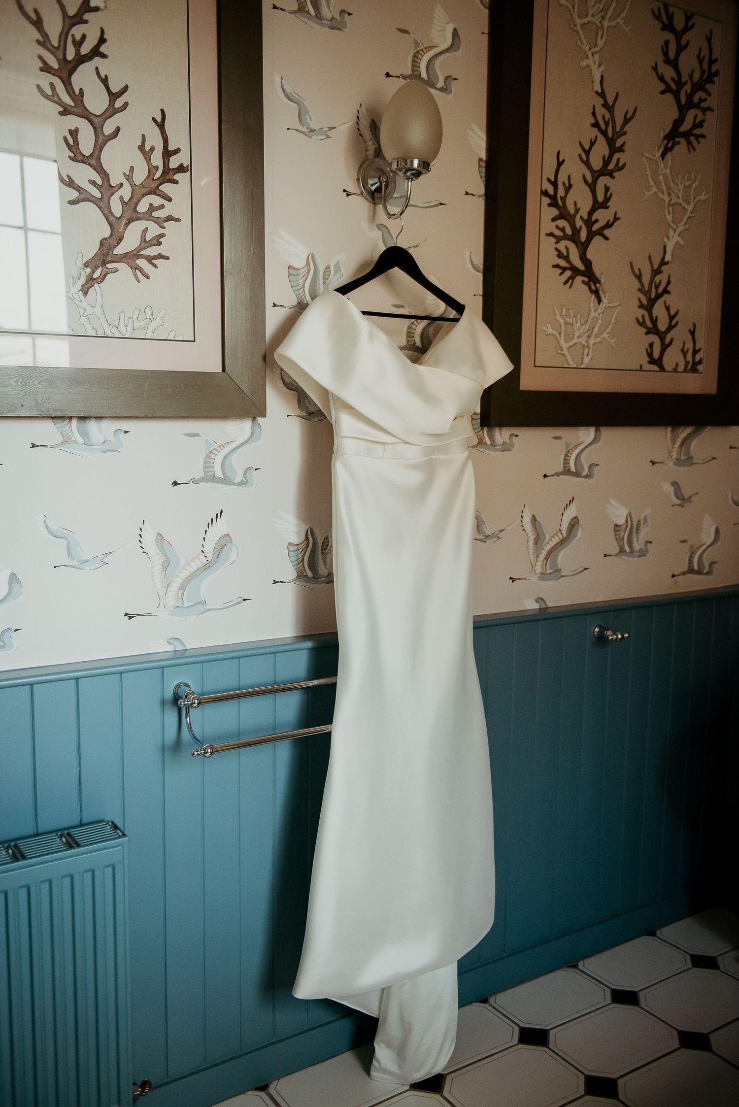 White wedding dress hangs on display against blue wainscoting and patterned wallpaper.