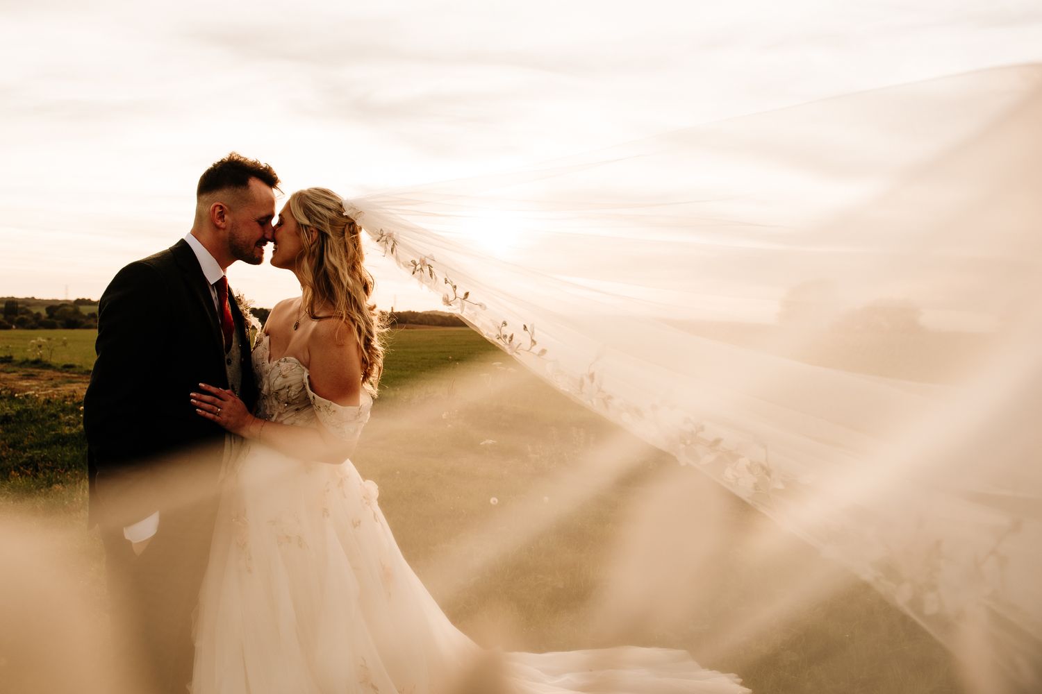 Romantic wedding couple kissing at sunset with bride's veil flowing in the wind in a scenic outdoor setting.