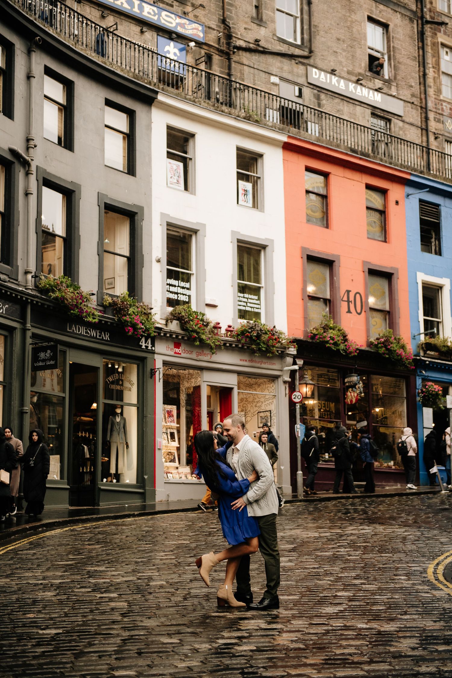 Couple embracing on cobblestone Victoria Street in Edinburgh with historic colorful buildings and shops in background.