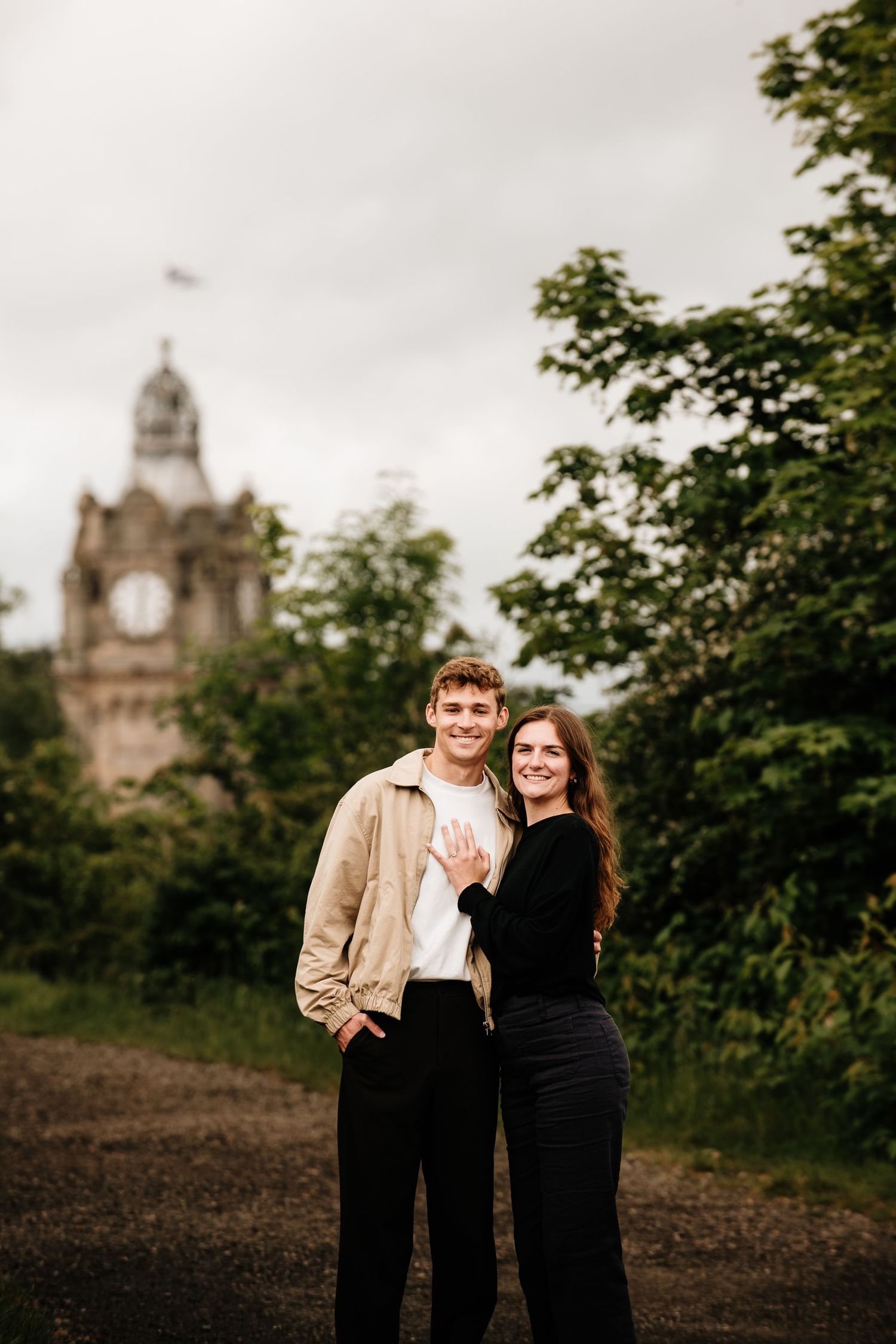 Happy couple posing outdoors near a historic clock tower surrounded by lush green trees on an overcast day.