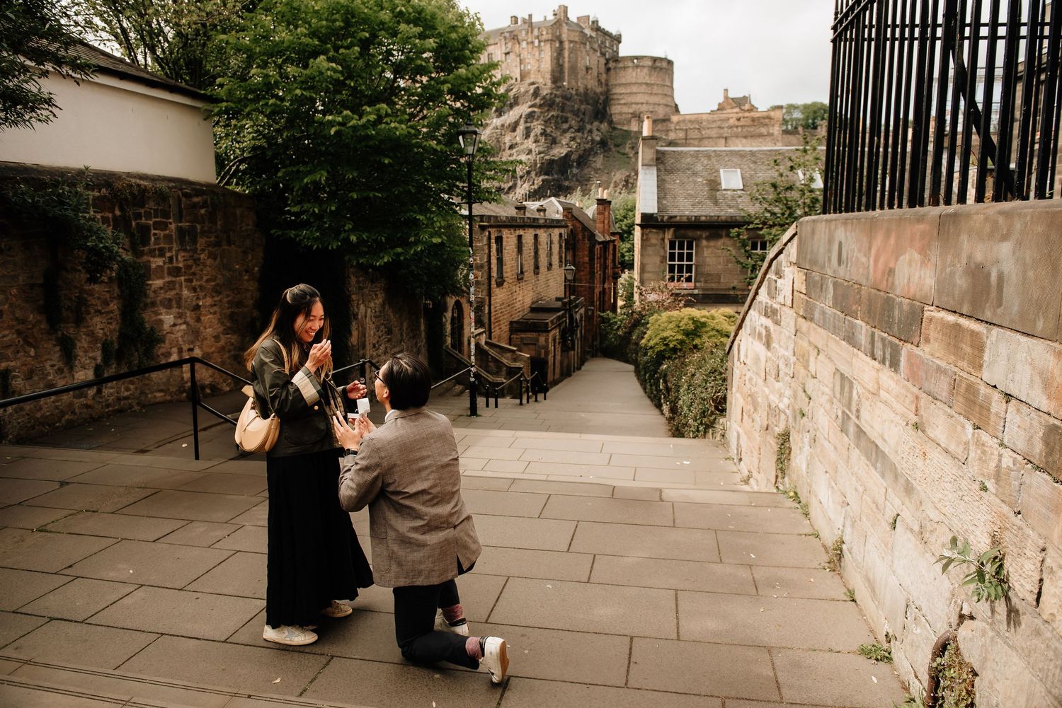 A surprise proposal moment unfolds on historic stone steps with Edinburgh Castle as backdrop.