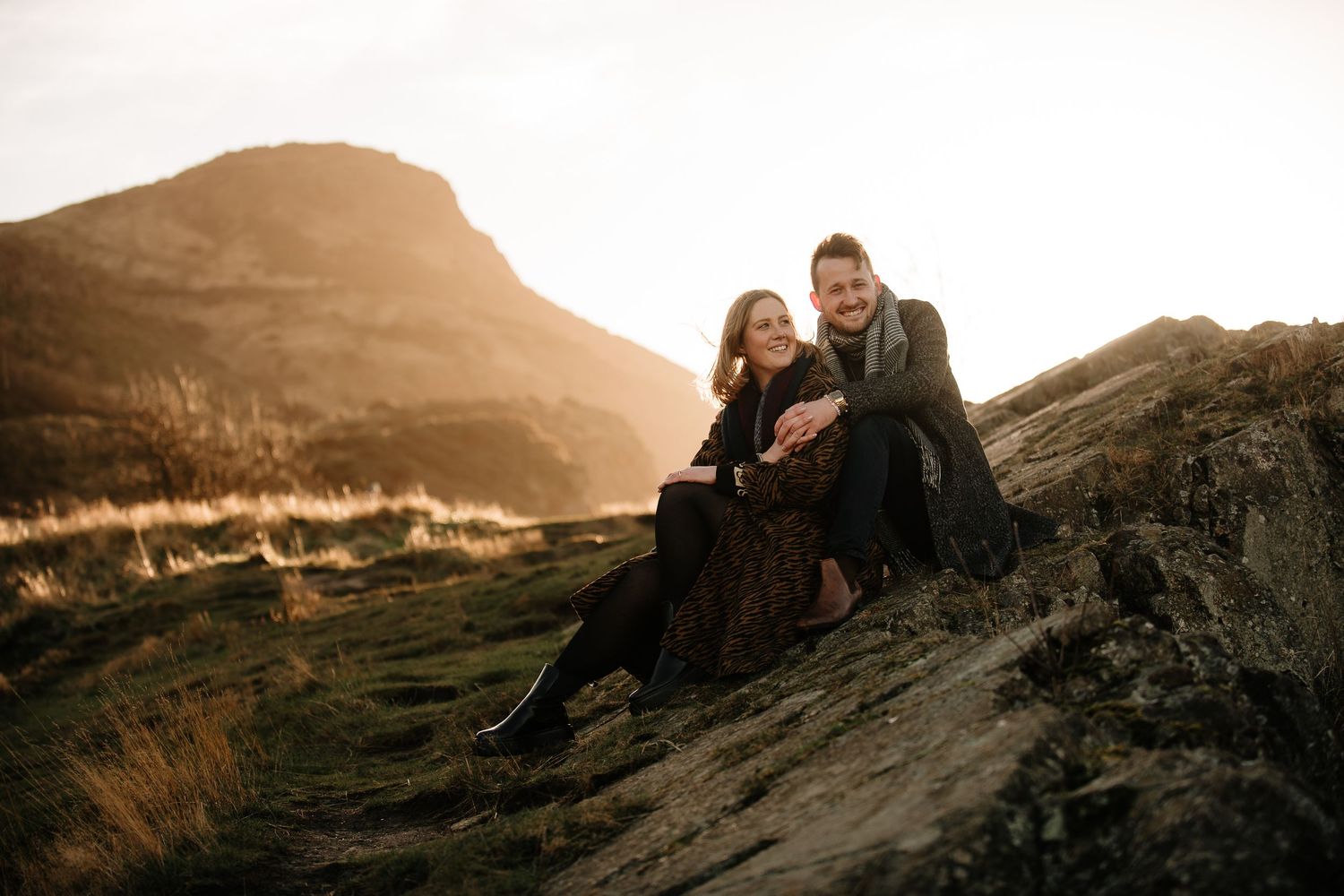 Couple sits together on rocky hillside during golden hour with mountain views.