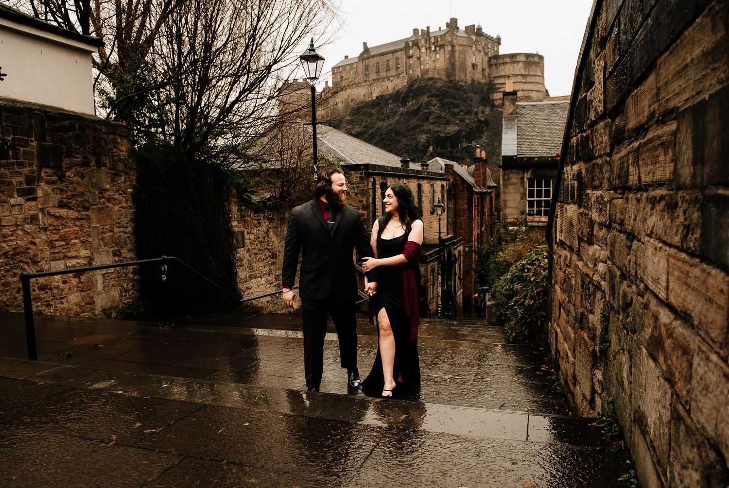 A couple stands in the rain on a cobblestone street with Edinburgh Castle looming in the background.