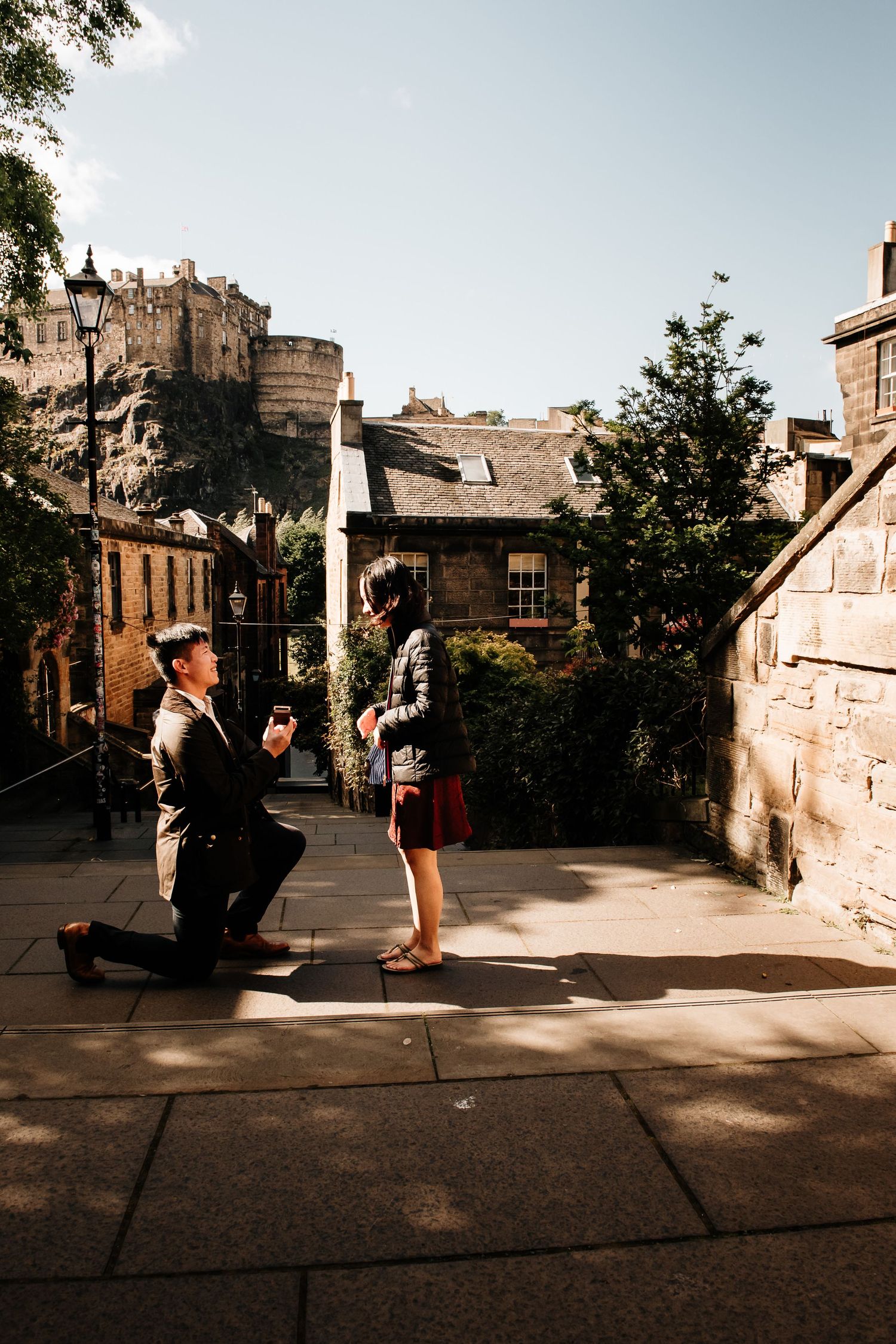 Man proposing on one knee to woman in sunny Edinburgh courtyard with castle rock backdrop.