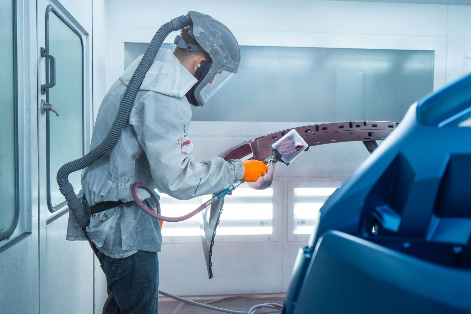 Auto body technician in protective suit and respirator painting a car panel in a professional spray booth.