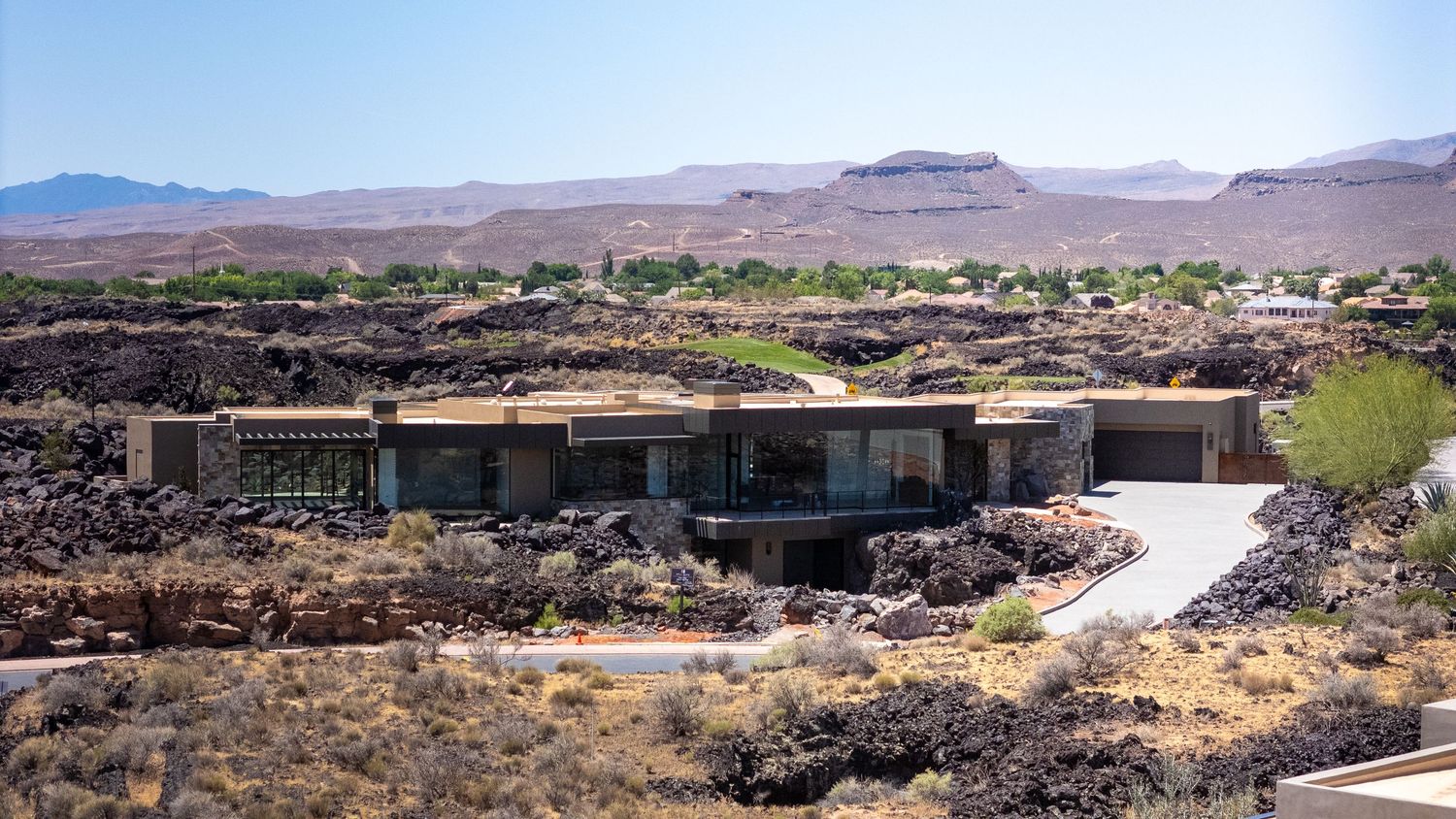Modern desert home with green roof, surrounded by volcanic rocks and mountain views under a clear blue sky.