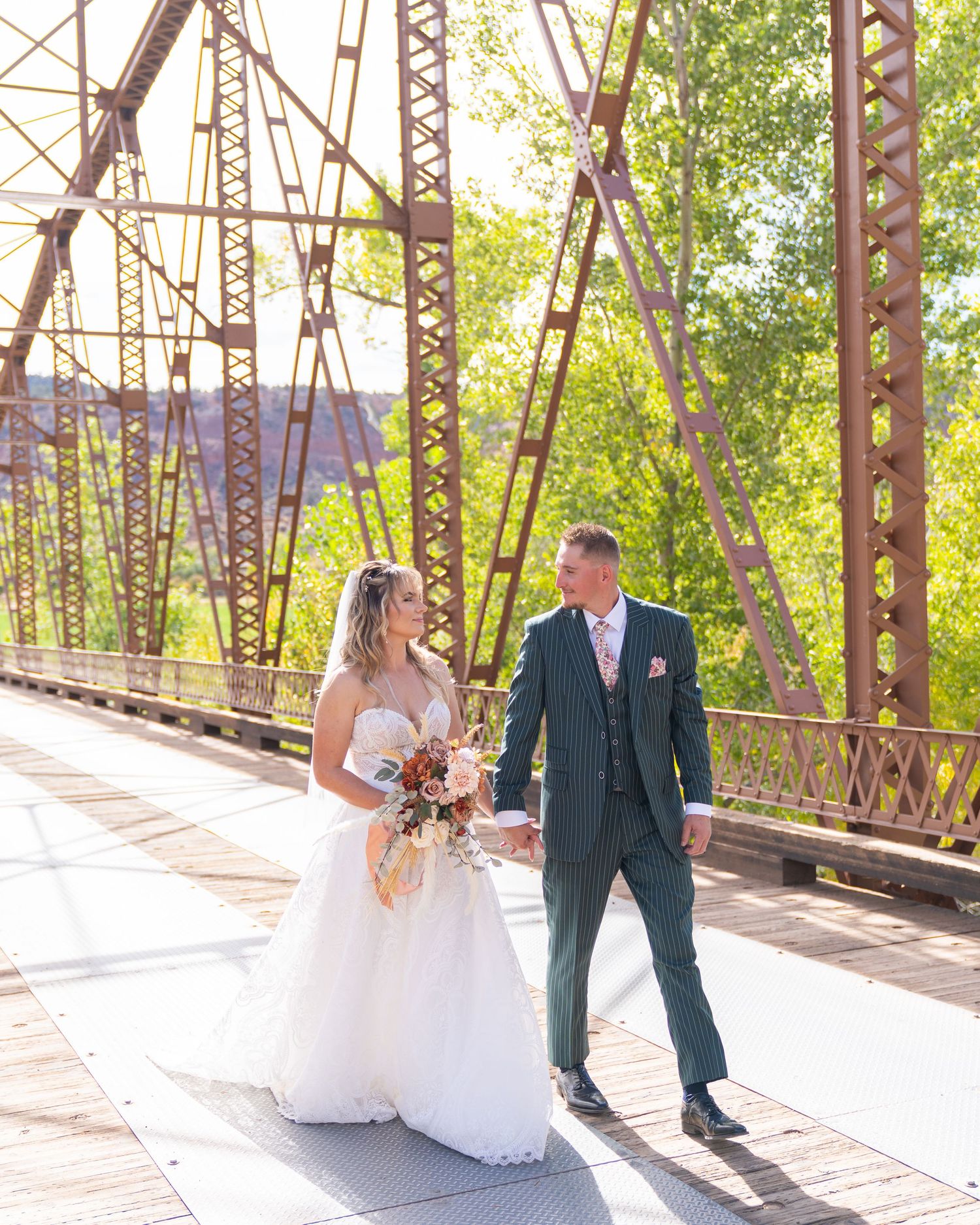 Bride and groom walk hand-in-hand on a rustic iron bridge, surrounded by lush green trees on their wedding day.