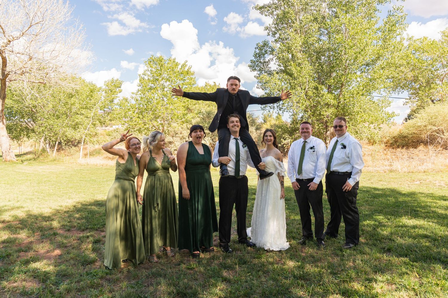 Wedding party poses outdoors on green lawn, bridesmaids in sage green dresses, groomsmen in suits, groom lifted above crowd.