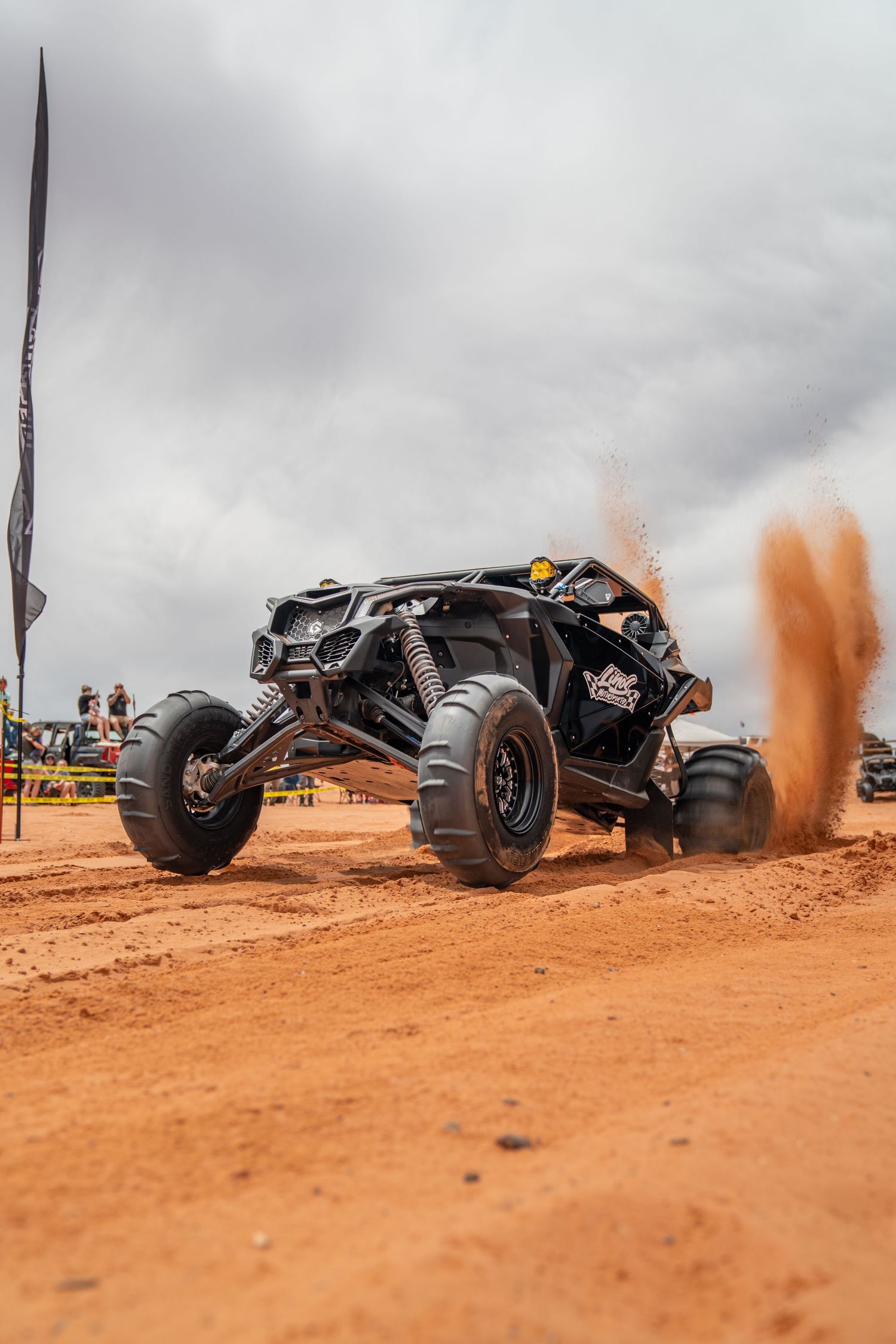 Black off-road UTV racing on red dirt track, kicking up dust clouds under overcast skies at outdoor motorsport event.