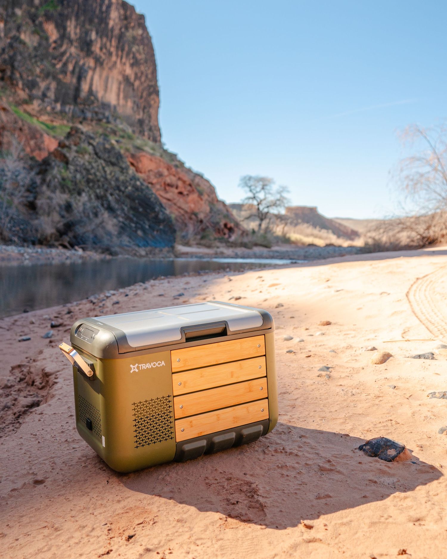 Portable olive green cooler with wood panel accents sitting on red sand beach beside a river with canyon cliffs in background.