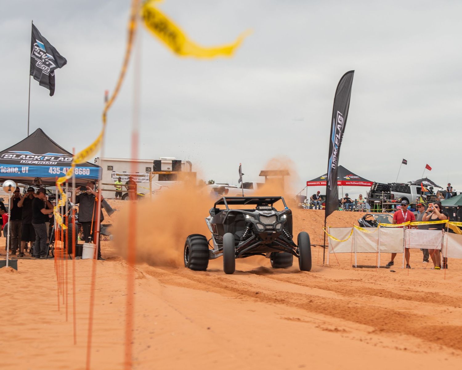A Can-Am UTV races through sandy terrain at an off-road event with sponsor flags and spectators in the background.
